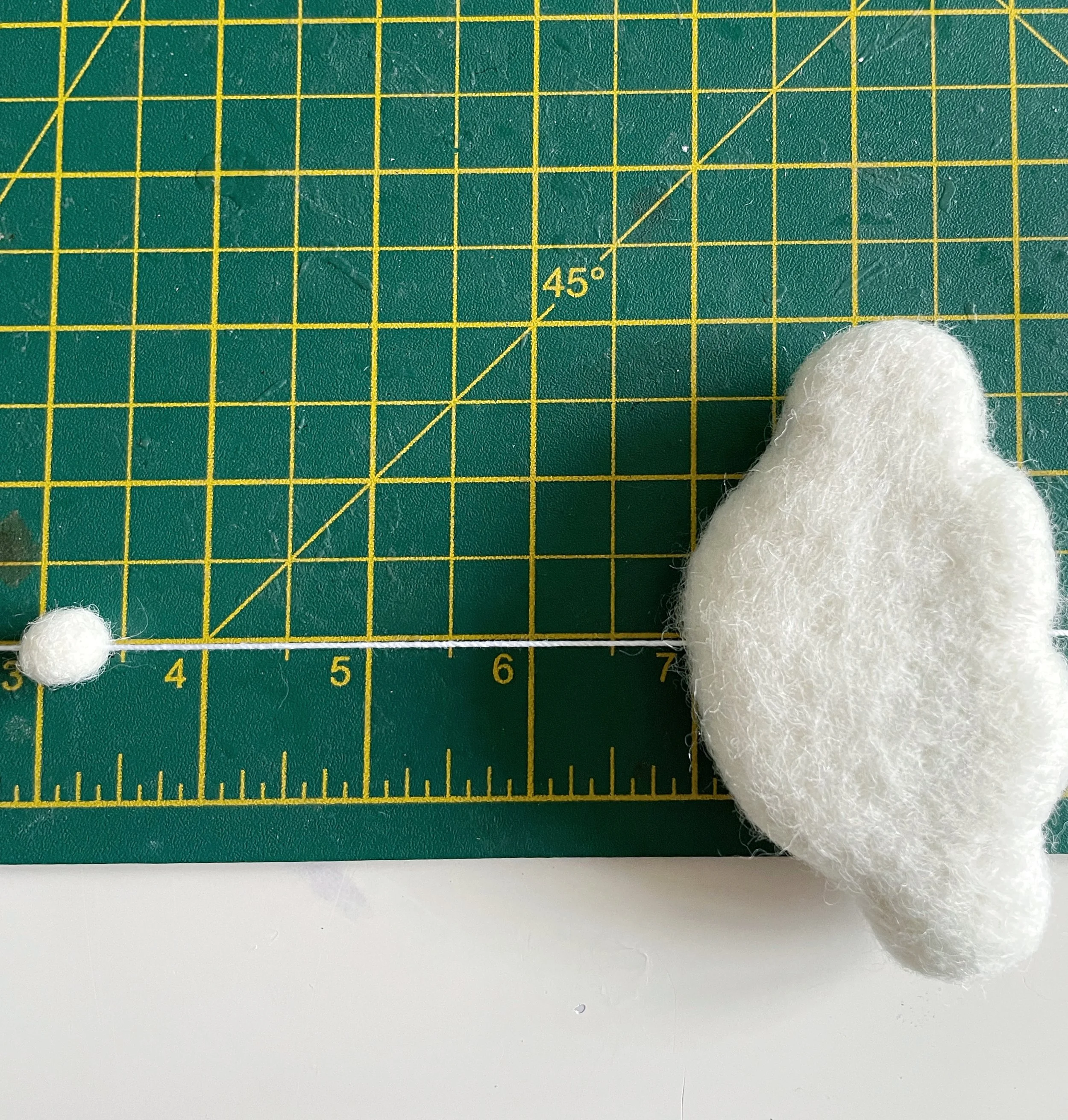 White string threaded with white felt ball and white wool felt cloud. Measured on a green cutting mat.