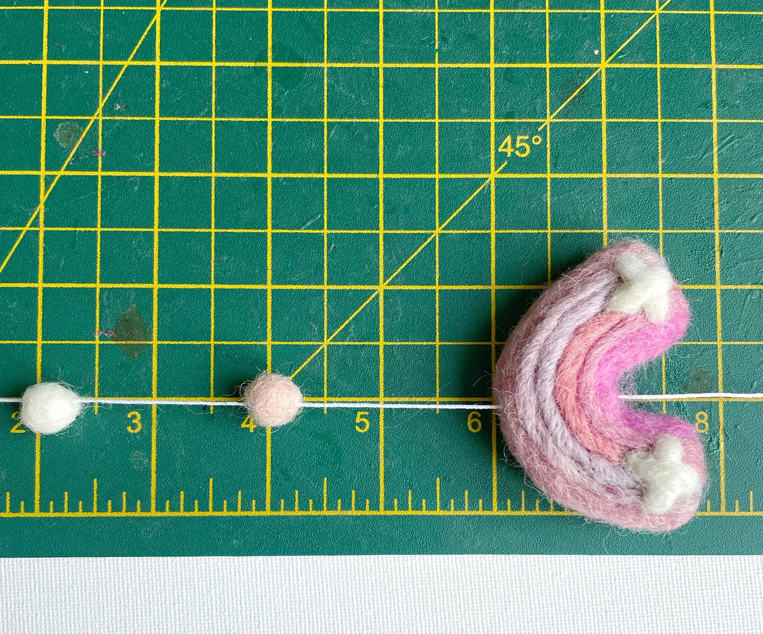 wool felt rainbow and a pink felt ball and a white felt ball, threaded on to white string, being measured on a green cutting mat.