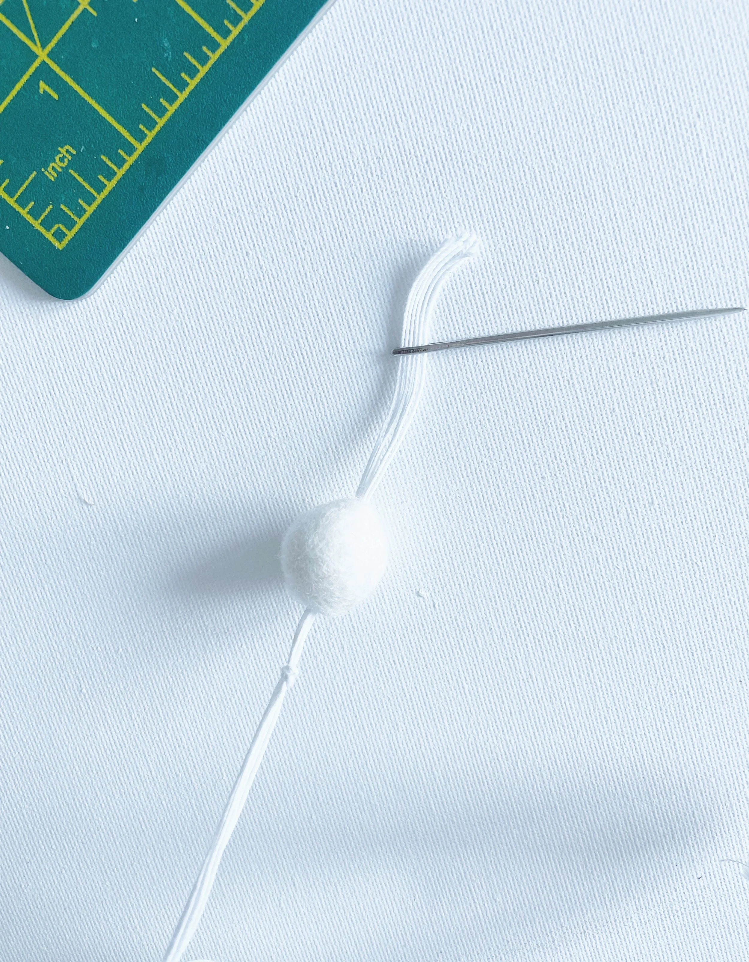 Six strands of white string threaded through sewing needle. White felt ball is threaded onto the  string.