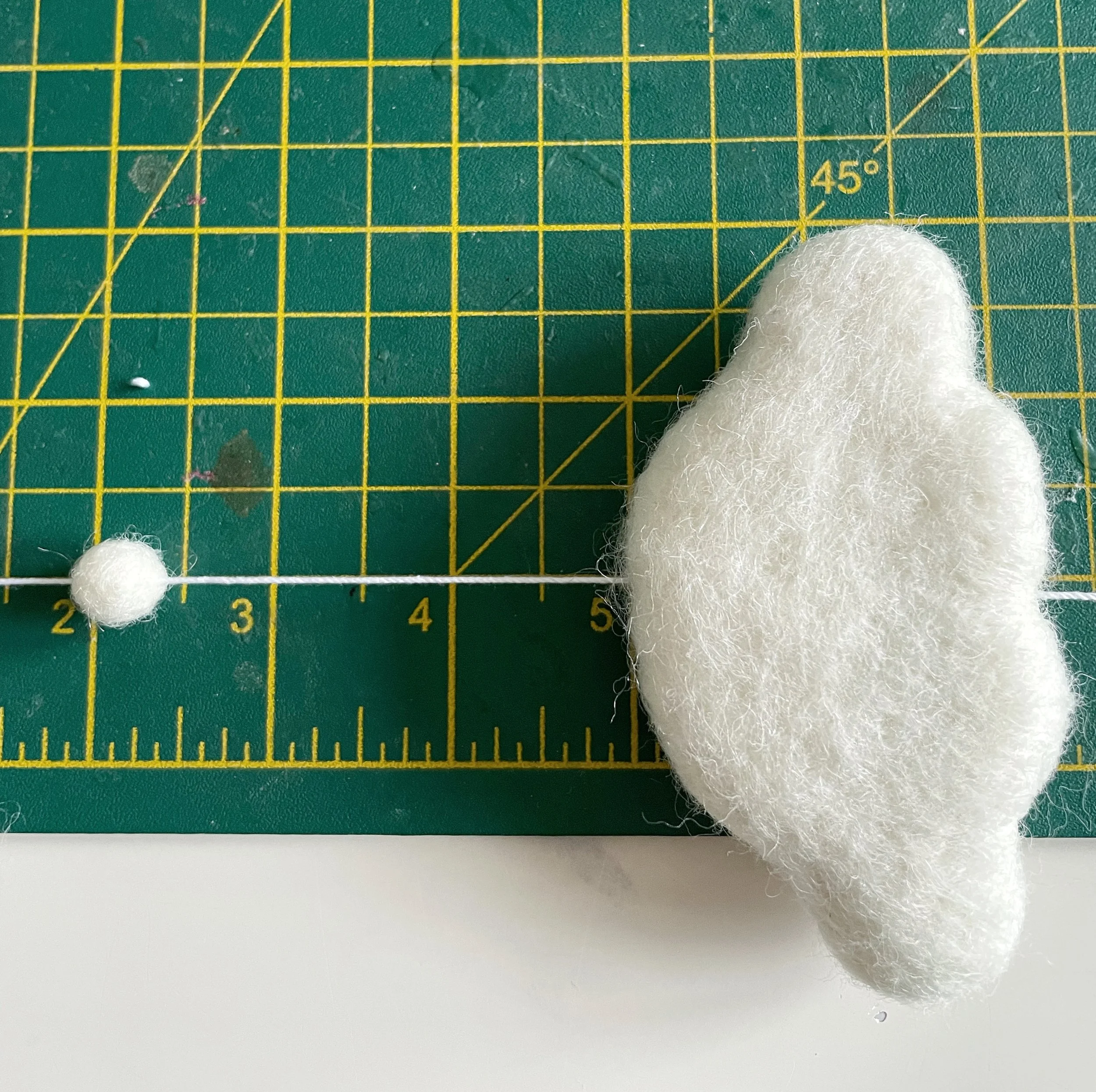 White string threaded with a white felt ball and a white cloud shape, measured on a green cutting mat.