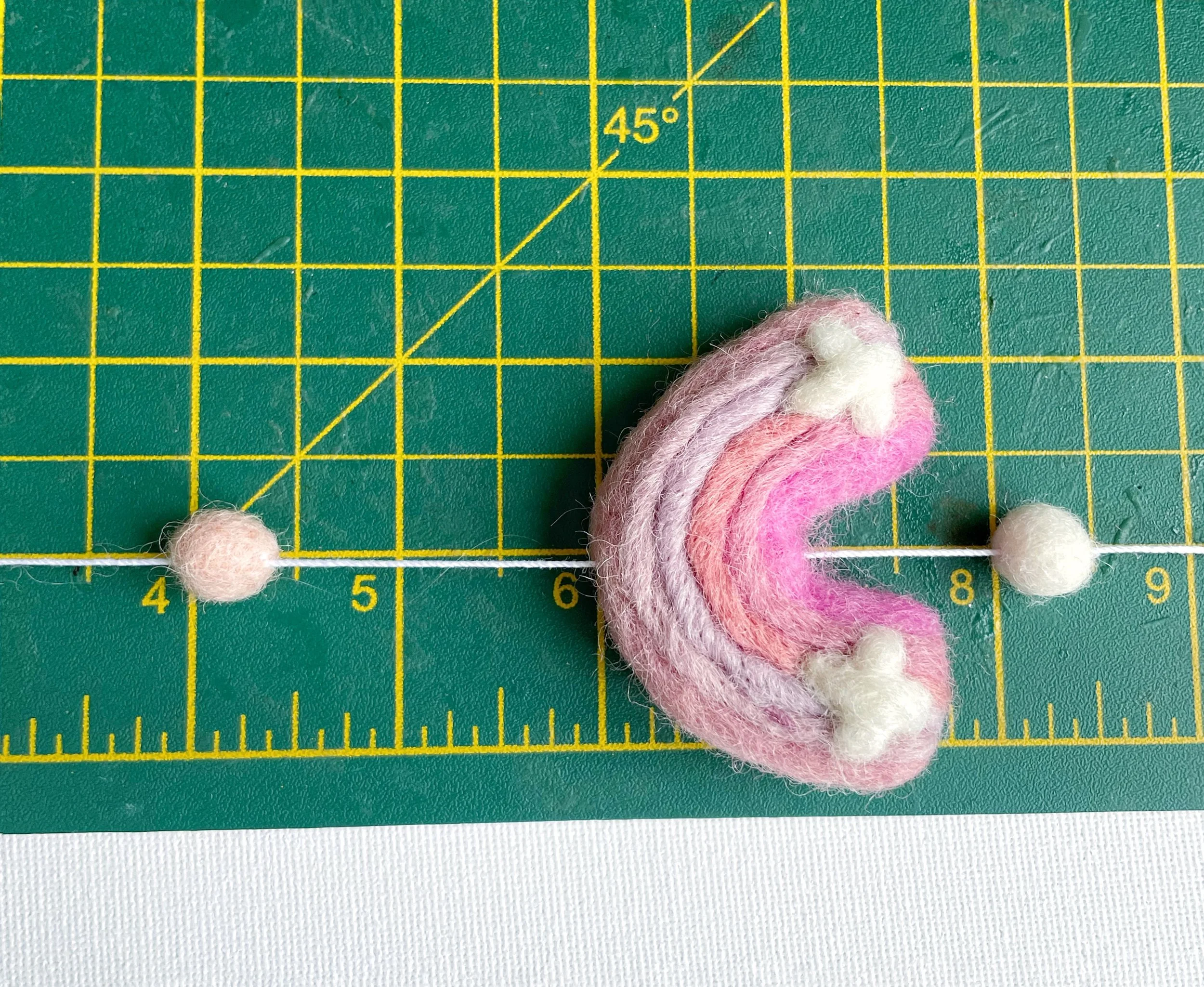 Wool felt rainbow, pink and white felt ball threaded onto white string. Measured on green cutting mat.