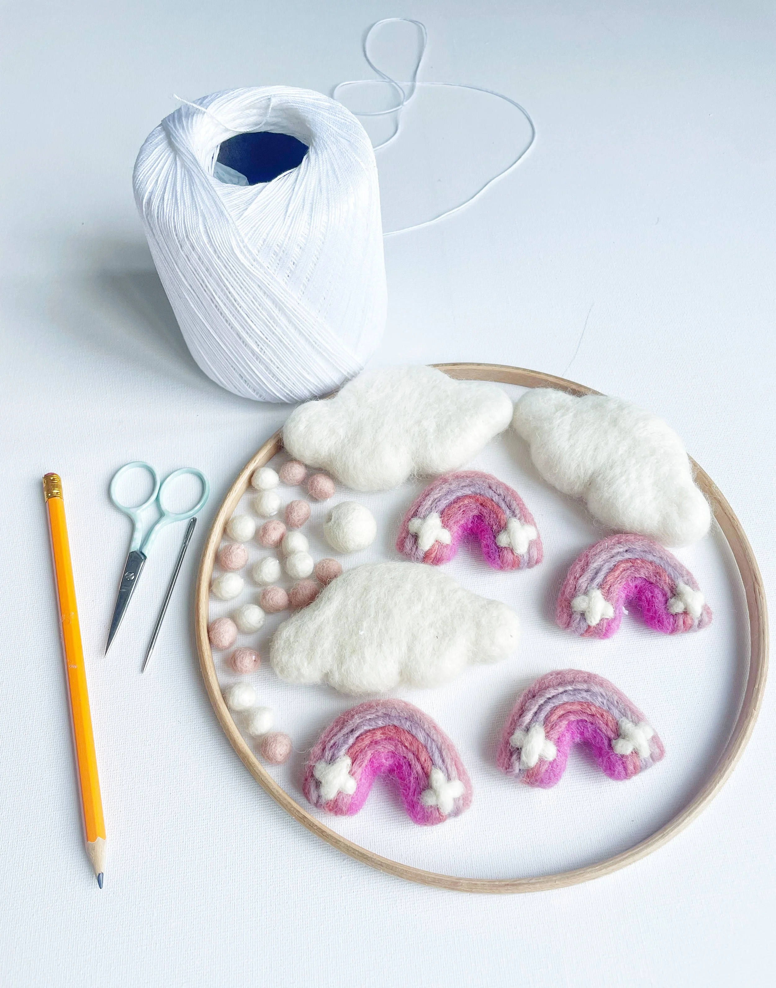 Reel of white string, wooden hoop, three white felt clouds, pink and white felt balls, small scissors, sewing needle and a pencil. Arranged on a white background.