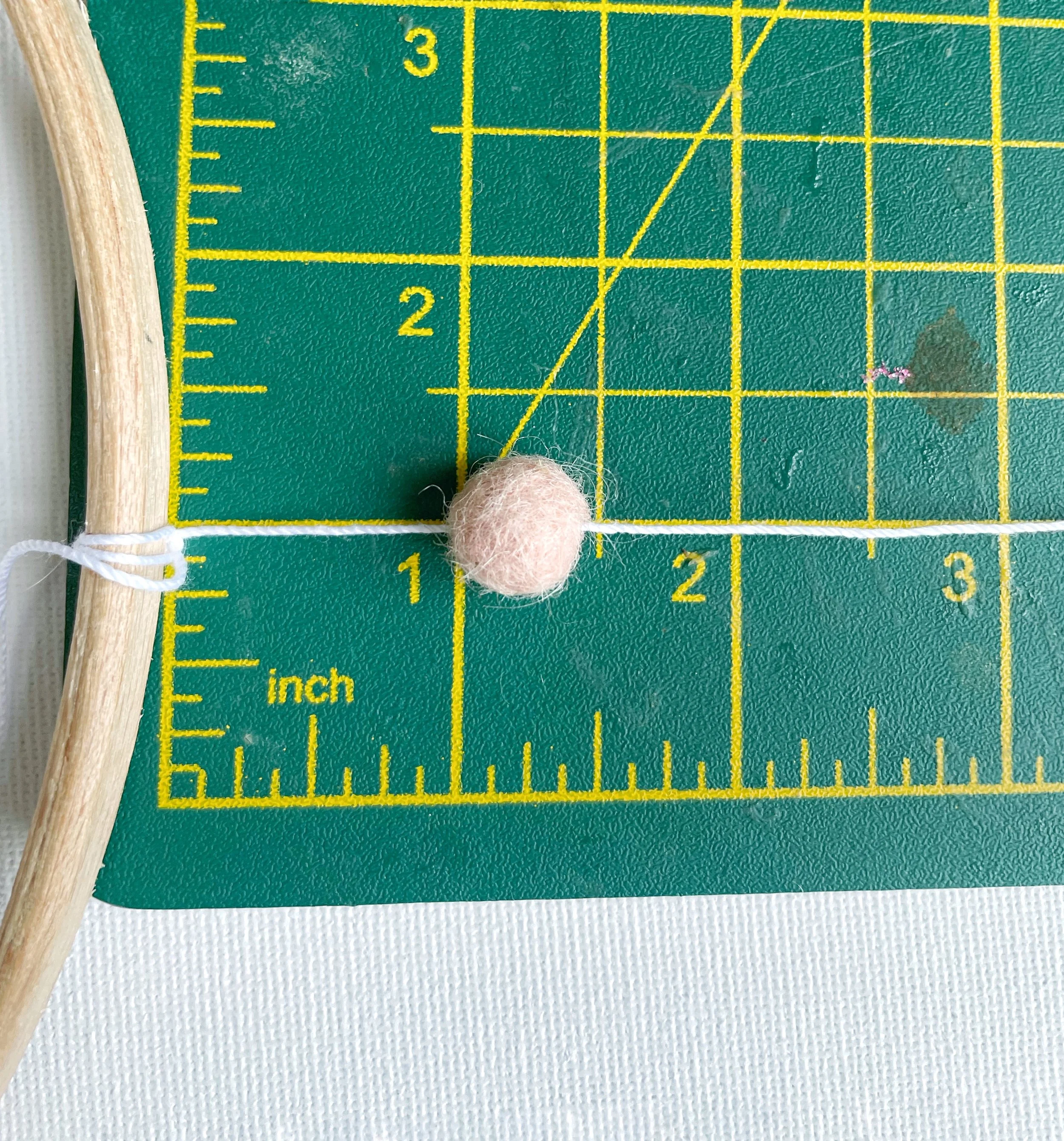 Pink felt ball threaded onto white string, attached to wooden hoop. Measured on green cutting mat.