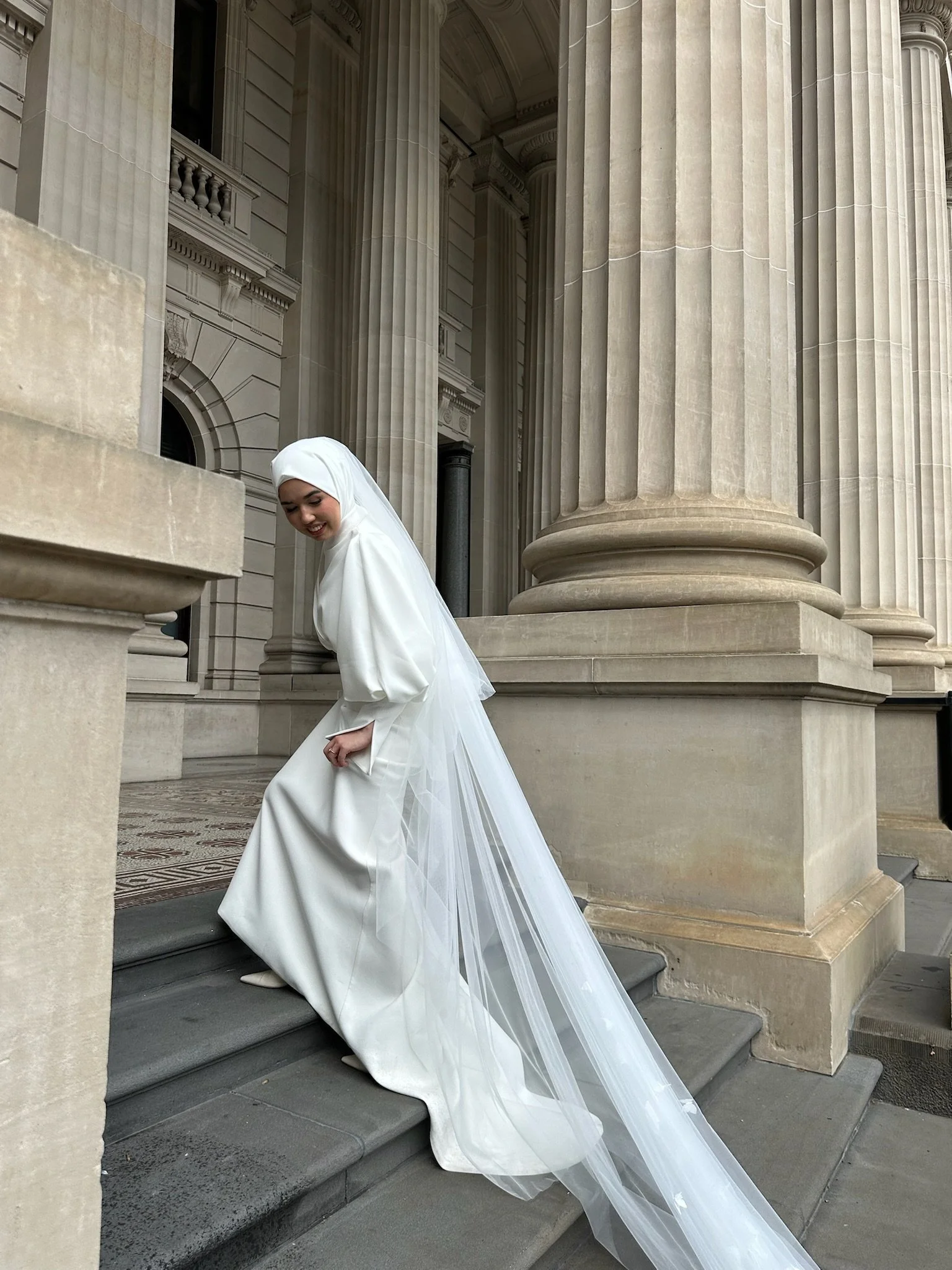 Woman in a white wedding dress and veil ascending steps near a classical architecture building with large columns. Captured by Melbourne Wedding Content Creator Dania.