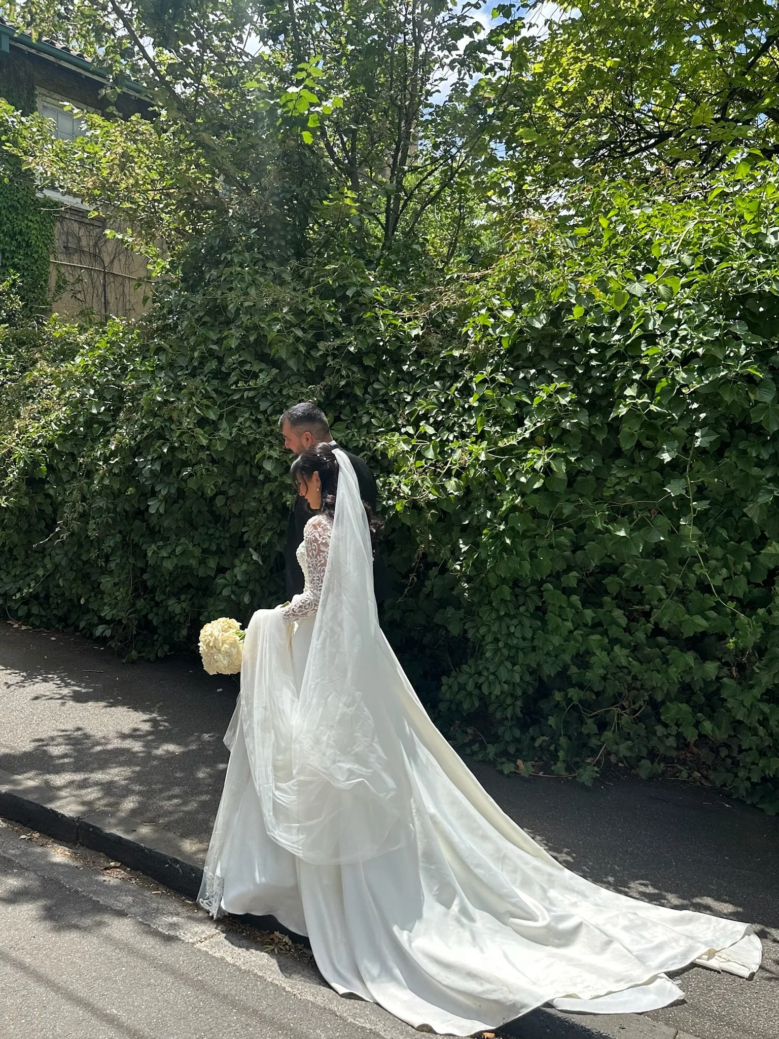 Bride in white wedding dress with train, holding bouquet, standing with groom in outdoor setting with greenery background. Captured by Melbourne Wedding Content Creator Dania.
