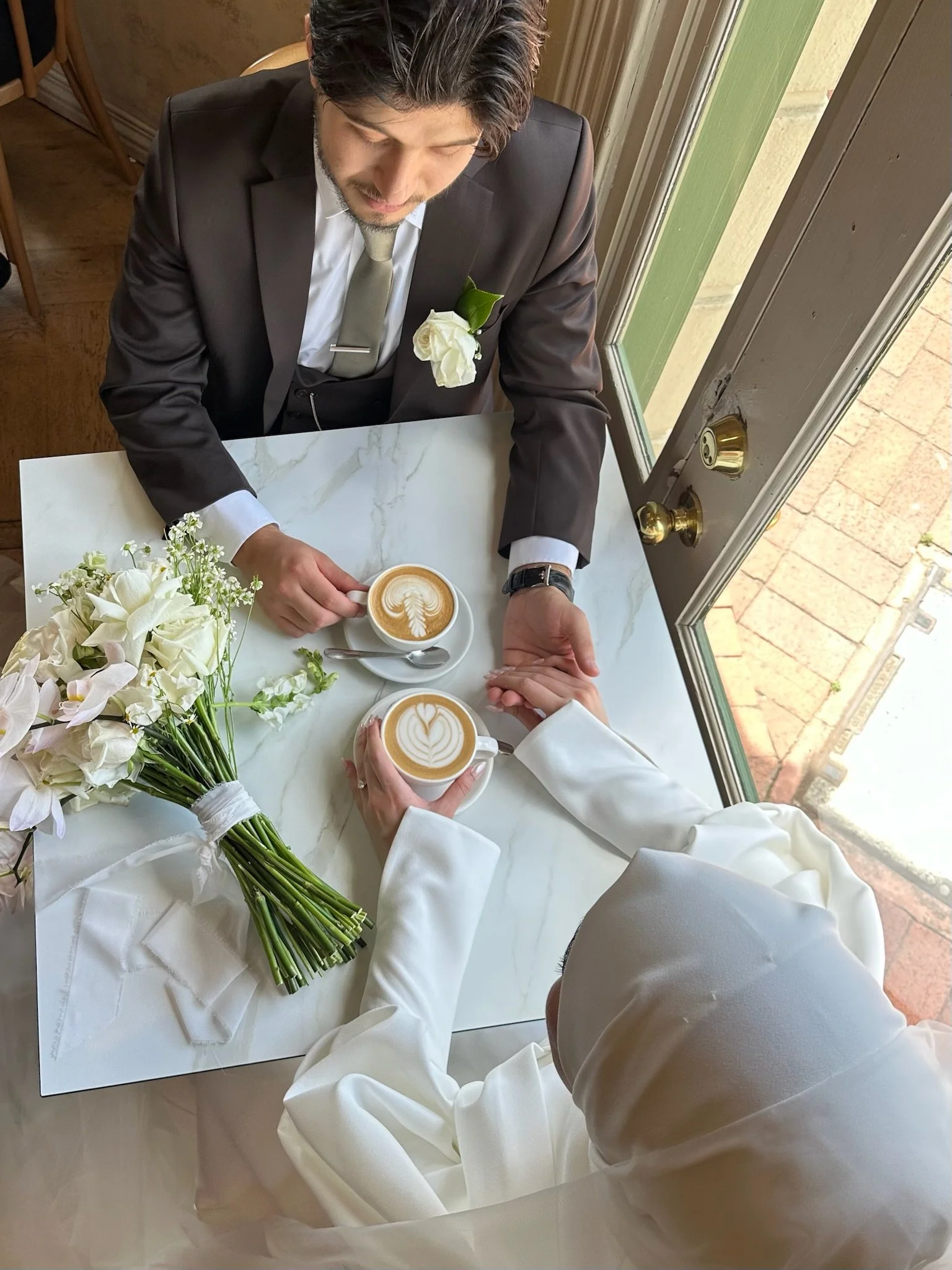 Couple in wedding attire holding hands with coffee at a table, bouquet present. Captured by Melbourne Wedding Content Creator Dania.