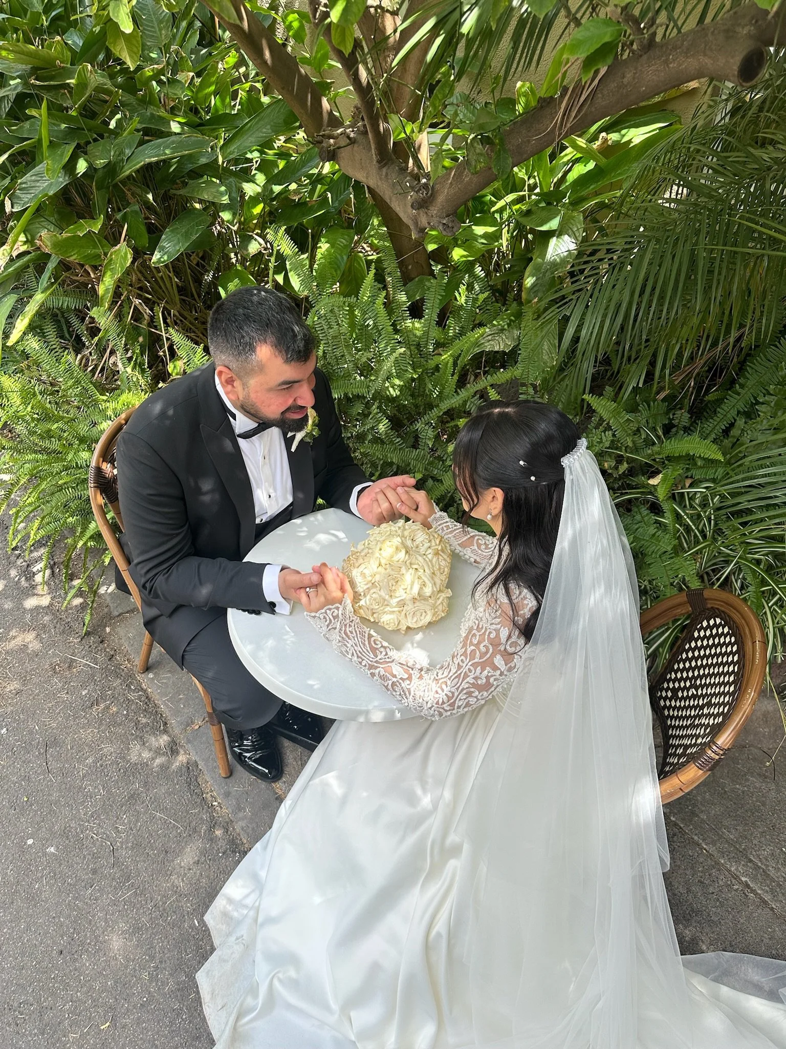 Bride and groom holding hands at a small table outdoors, surrounded by lush greenery, with the bride's bouquet on the table. Captured by Melbourne Wedding Content Creator Dania.