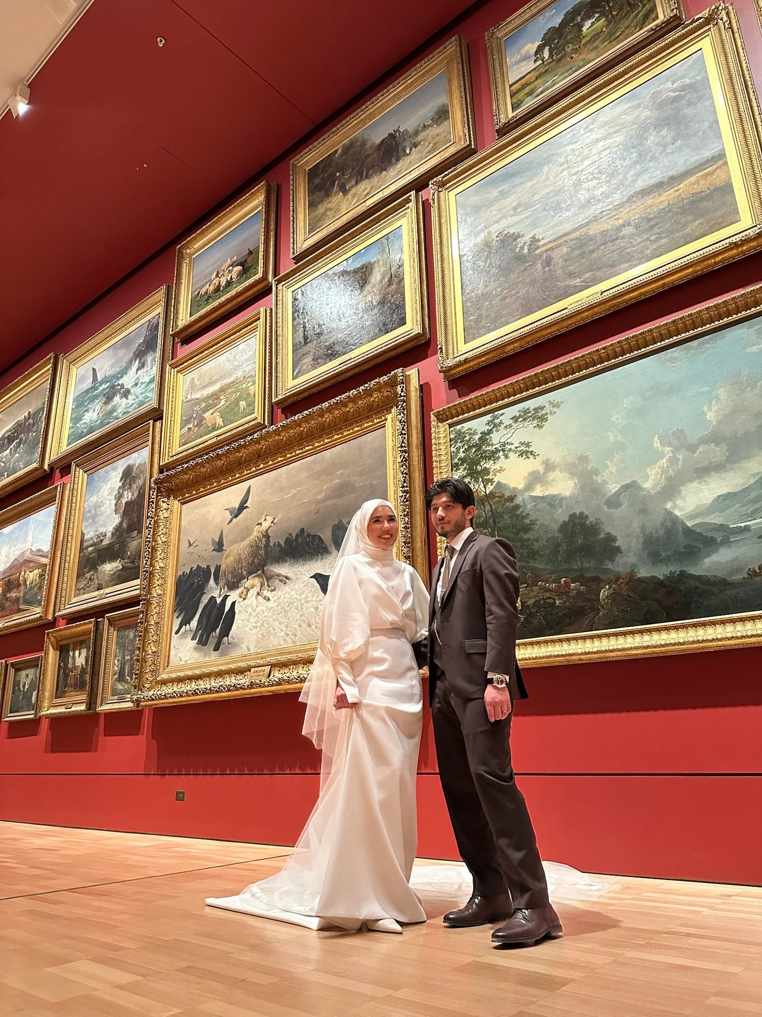 A couple in formal attire stands in an art gallery with red walls, surrounded by large framed paintings. Captured by Melbourne Wedding Content Creator Dania.