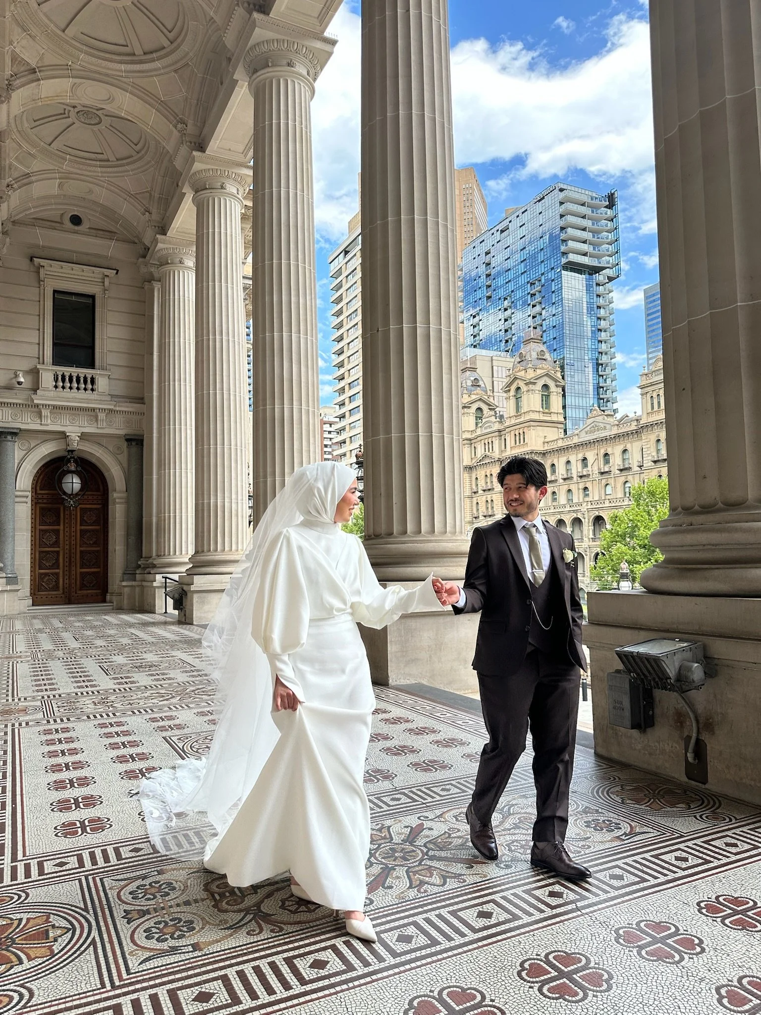 Wedding couple walking under a columned portico, with cityscape in the background. Captured by Melbourne Wedding Content Creator Dania.