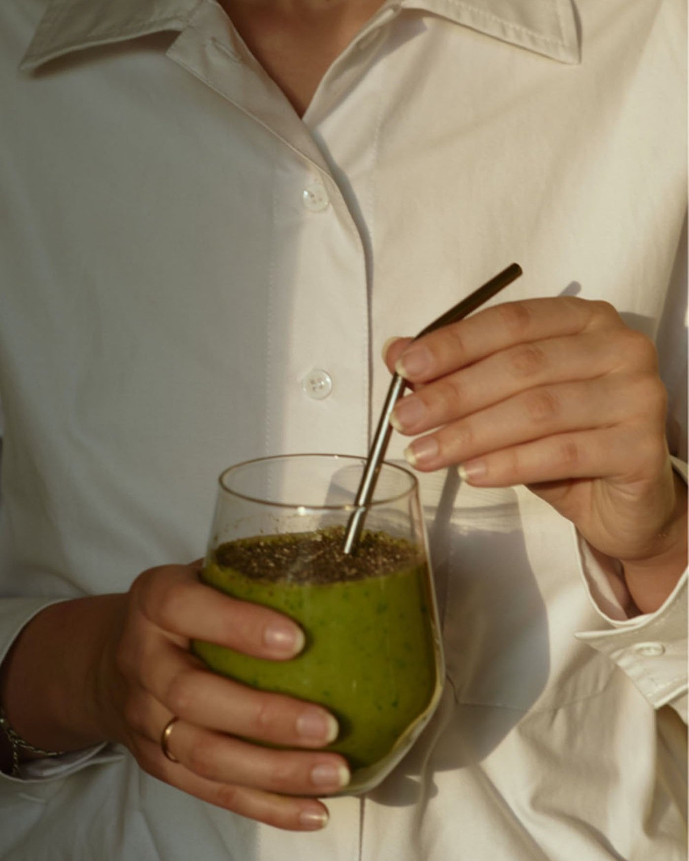 Person in a white shirt stirring a green smoothie in a glass with a metal straw.