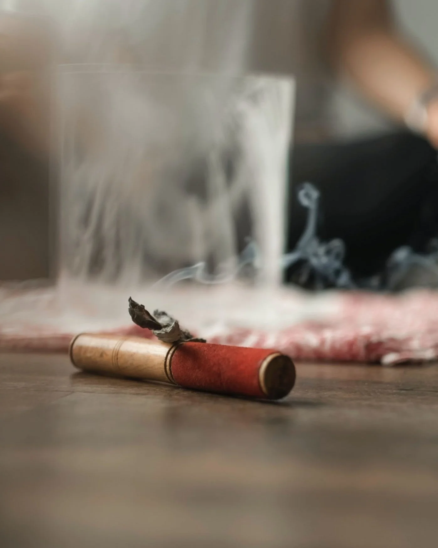 A partially burnt cigarette lying on a wooden surface with a grayish ash and smoke, blurred background including a glass and a black object.