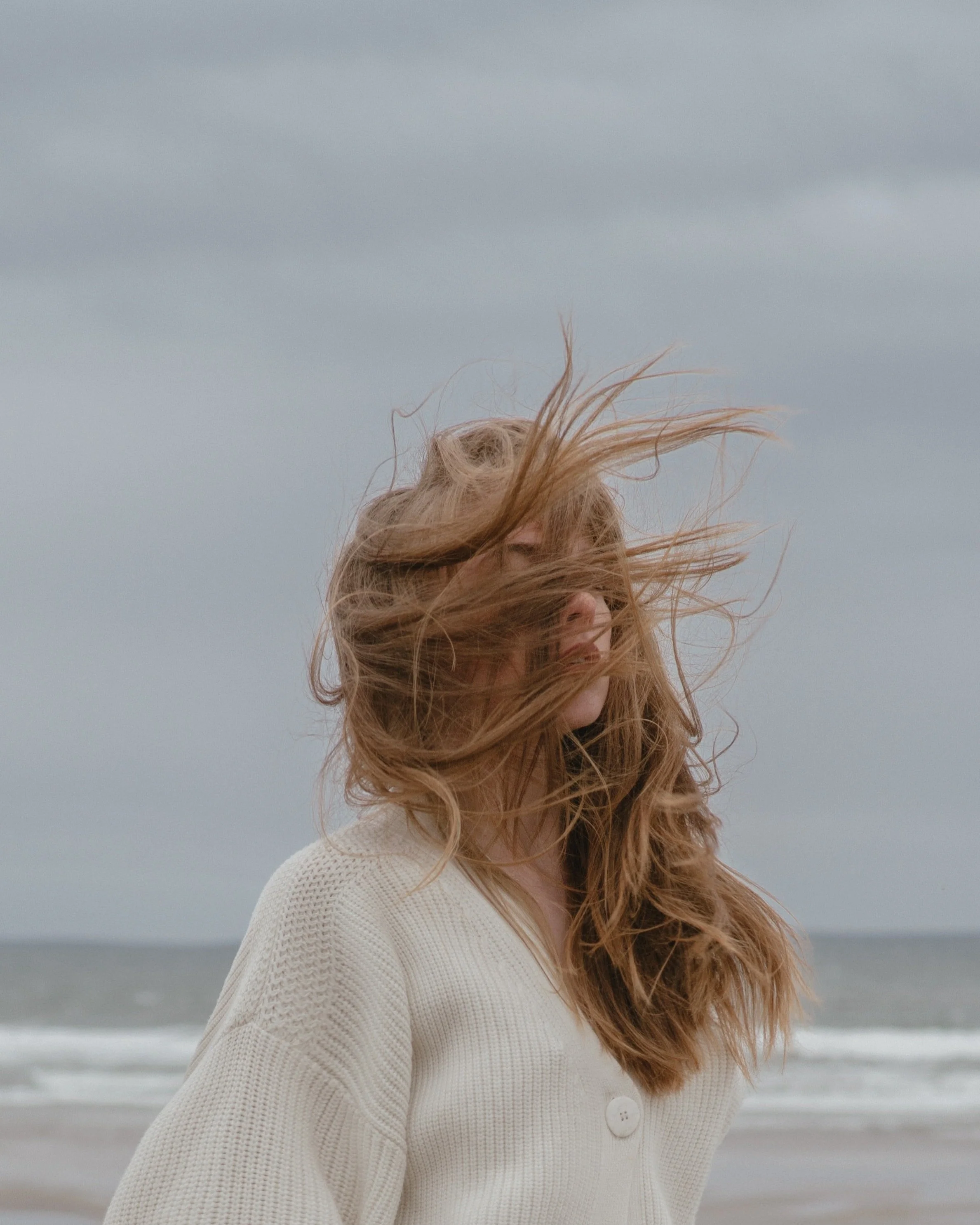 A woman with long, wavy hair standing on a beach with gray clouds and ocean waves in the background, her hair blowing in the wind.