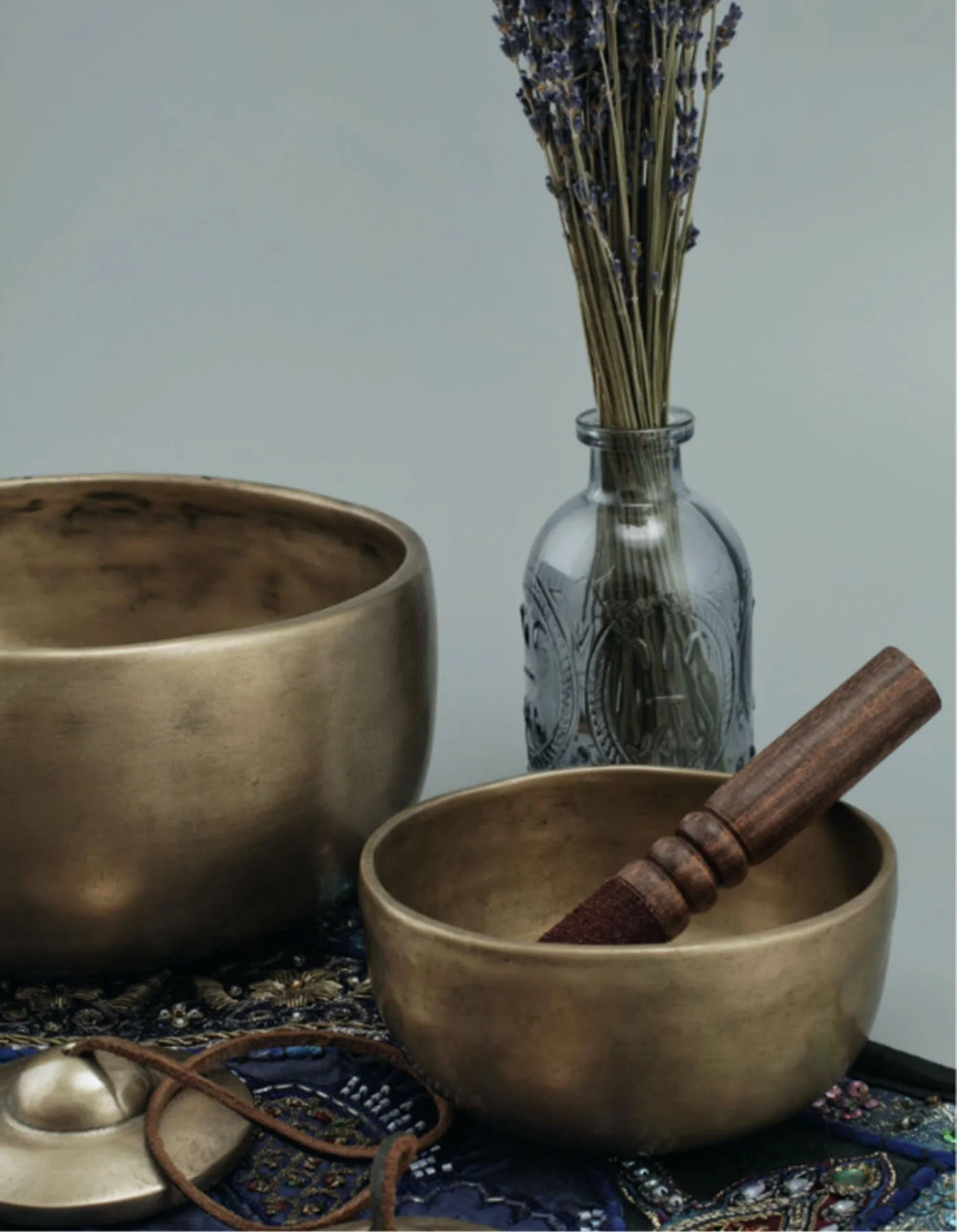 Two brass singing bowls, a glass vase with lavender flowers, and a wooden mallet, arranged on embroidered fabric.