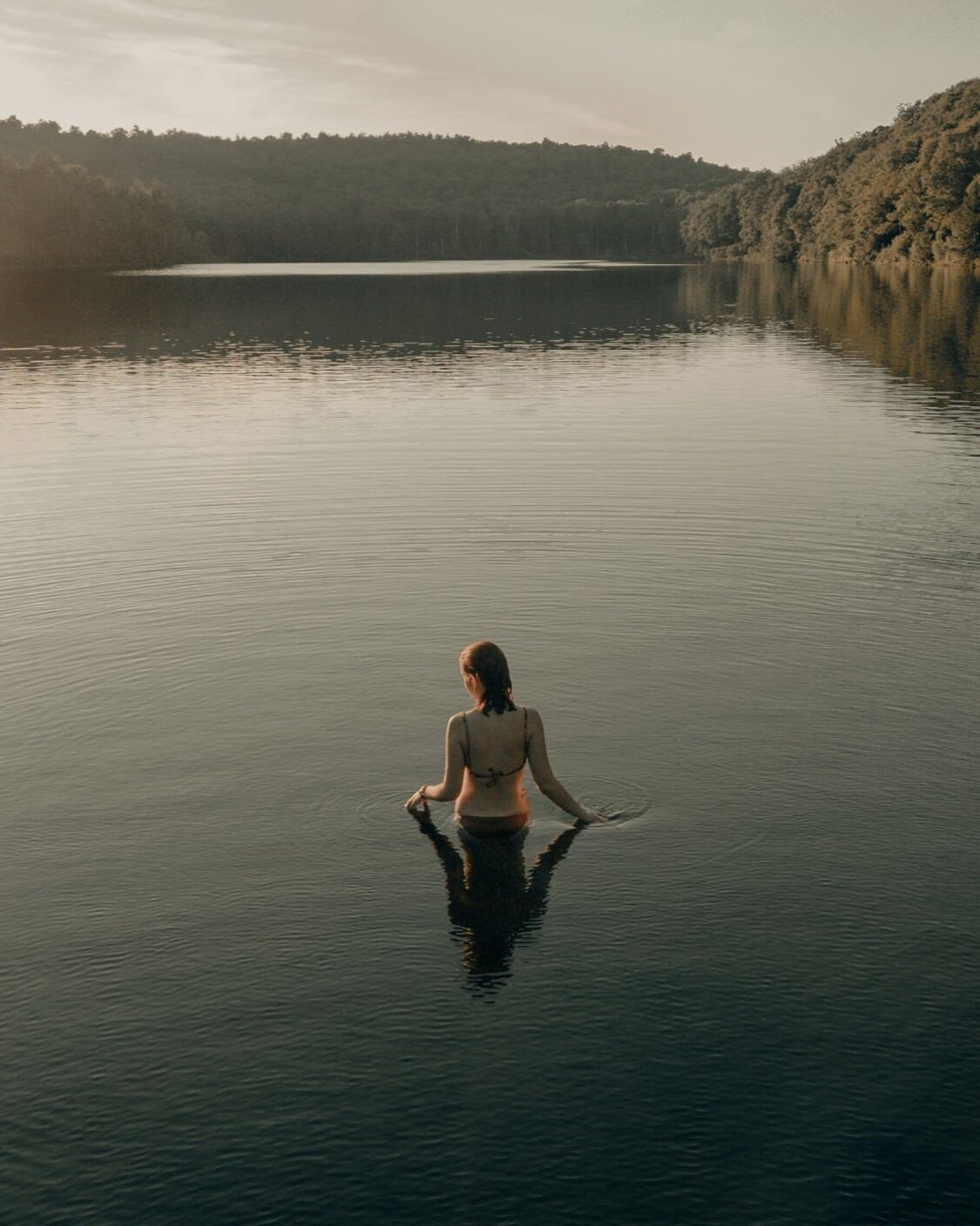 A woman in a swimsuit standing waist-deep in a calm lake, with a scenic view of hills and trees in the background during sunset or sunrise.