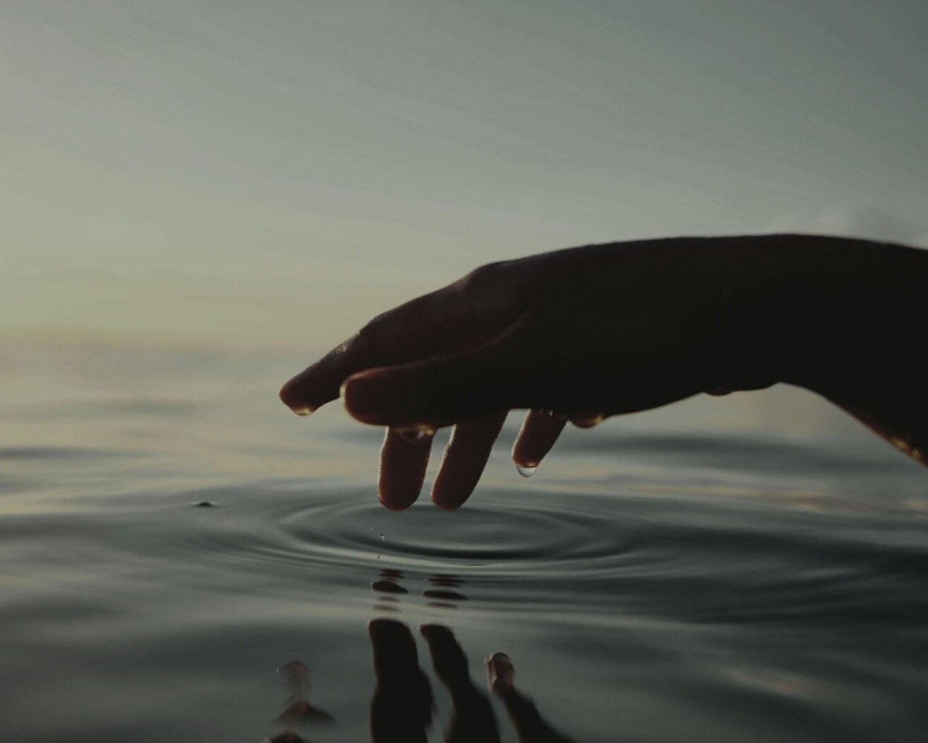 A person's hand skims the surface of a body of water, creating ripples, during sunset or sunrise with a calm sky in the background.