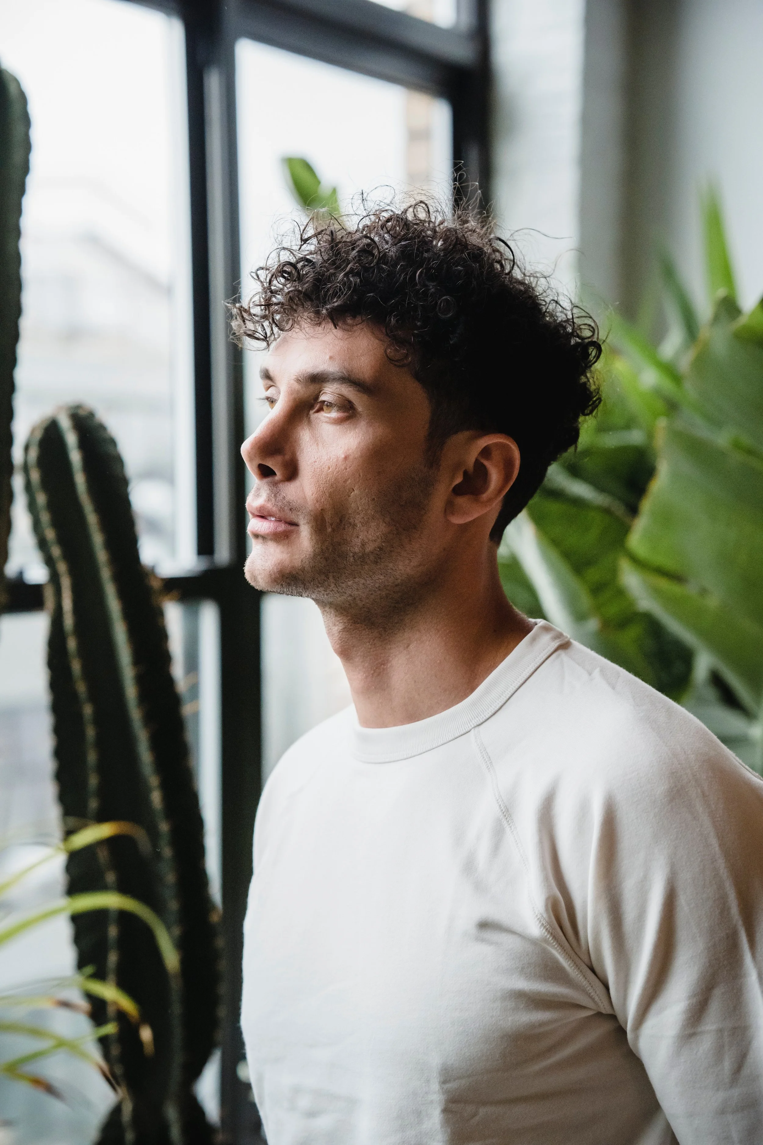 A young man with curly dark hair and light skin, wearing a white shirt, looking out a large window with green plants nearby inside.