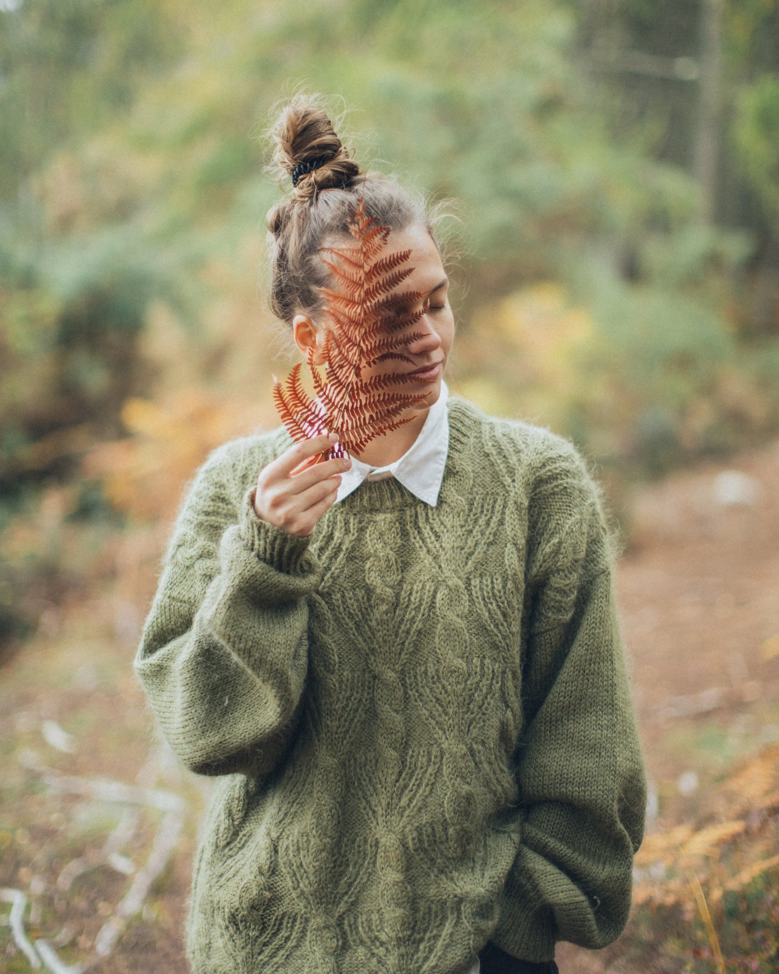 A woman with a messy bun hairstyle holding a reddish-brown fern leaf in front of her face with eyes closed, wearing a green knit sweater over a white collared shirt, standing outdoors with autumn foliage in the background.