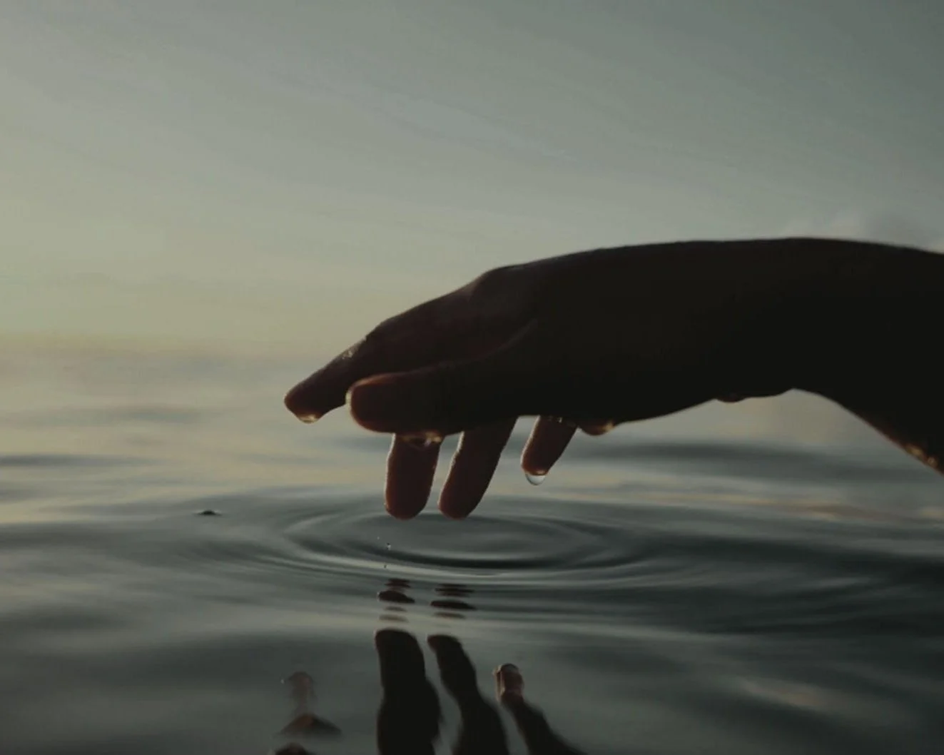 A person's hand reaching into a calm body of water with a rippling effect, against a backdrop of a sky at dusk or dawn.