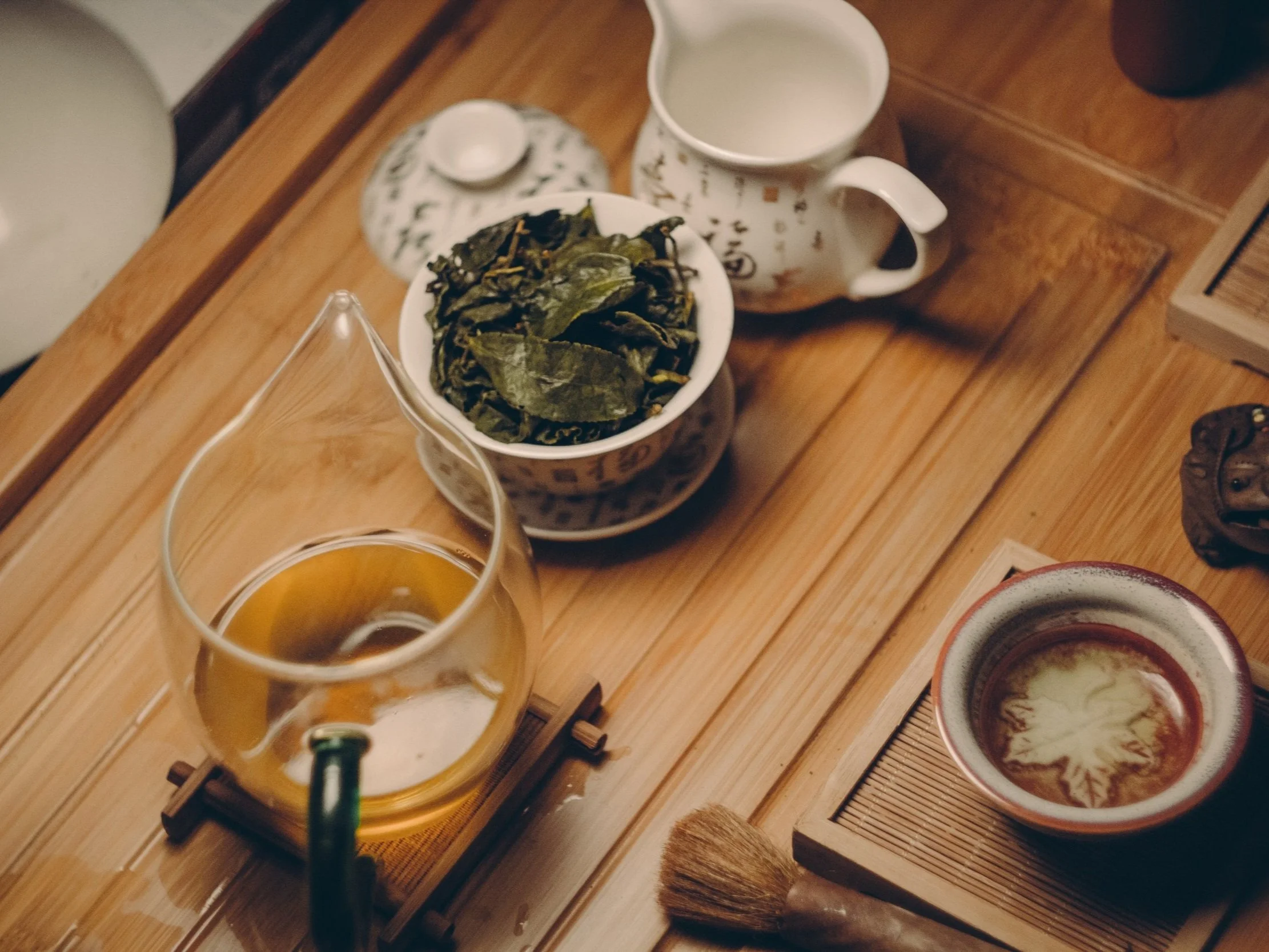 A tea setup on a wooden table includes a glass teapot with tea, a bowl of loose tea leaves, a cup with tea, a mug of milk, and a small bowl of brewed tea with intricate leaf patterns.