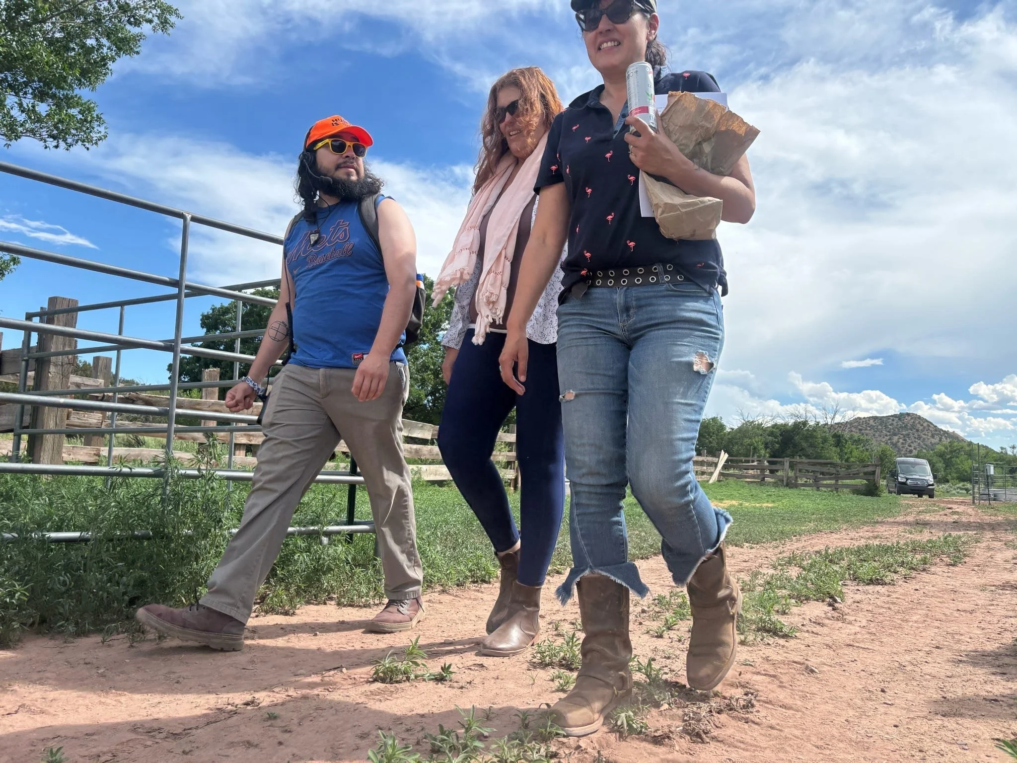 Three people walking outdoors on a dirt path, with a wooden fence and green trees in the background. The sky is partly cloudy. One person is wearing a blue tank top and beige pants, another a black shirt with pink flamingos and ripped jeans, and the third a pink scarf and dark pants.