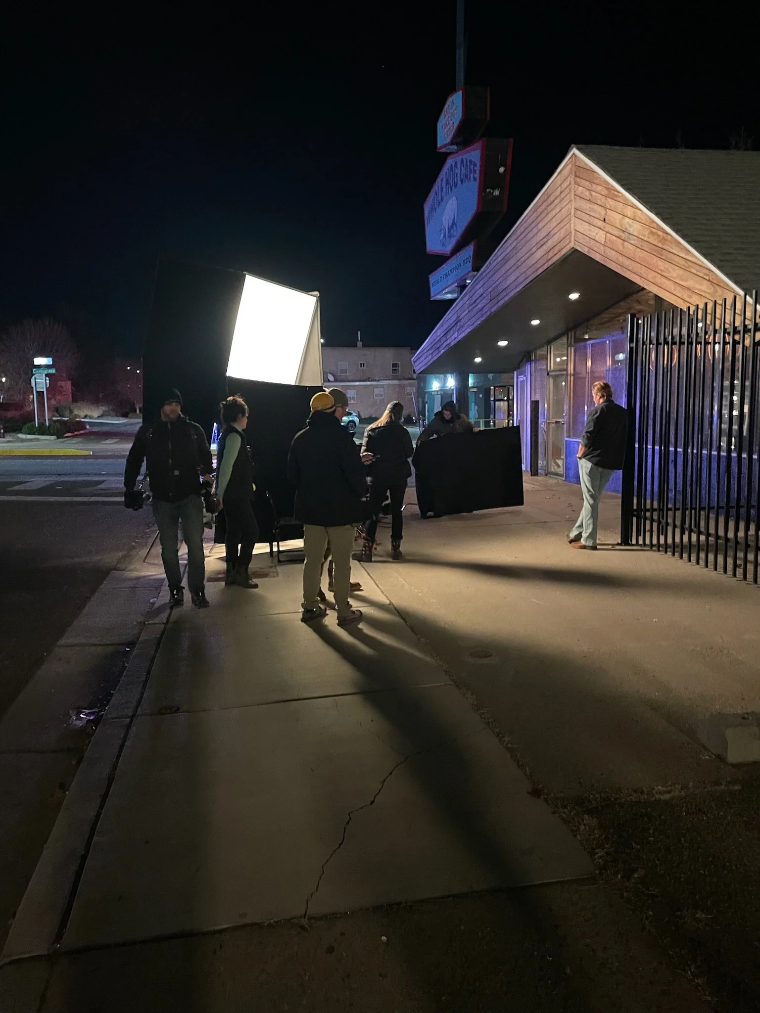Group of people standing outside a restaurant at night, some near a filming or photography setup with lighting equipment, near a building with a wooden exterior.