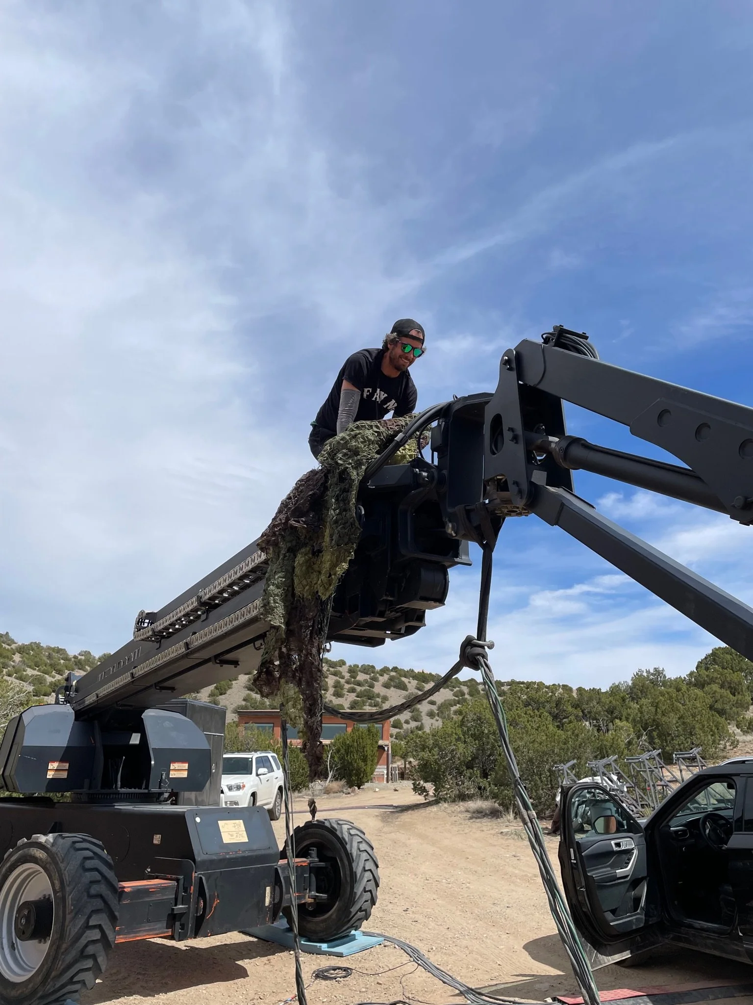 A man with sunglasses and a cap stands in a work platform attached to a wheel loader, with a camouflage cloth hanging from the platform. The setting is outdoors with a dirt road, a white vehicle, a van, and a hillside with trees in the background.