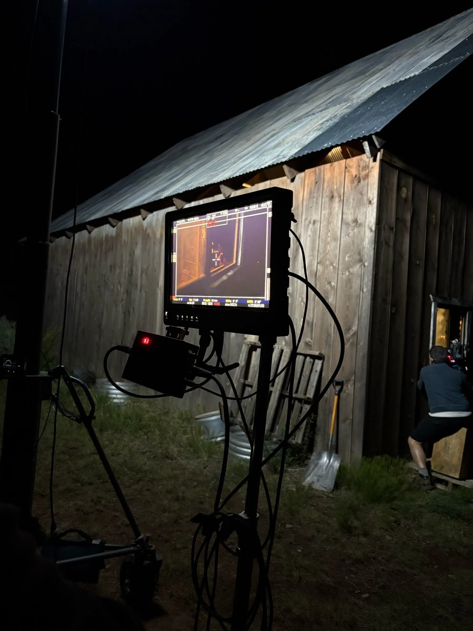 Nighttime scene with film equipment set up outside a wooden building, a monitor to the left displaying footage, and a person in shorts and a jacket working near the building.