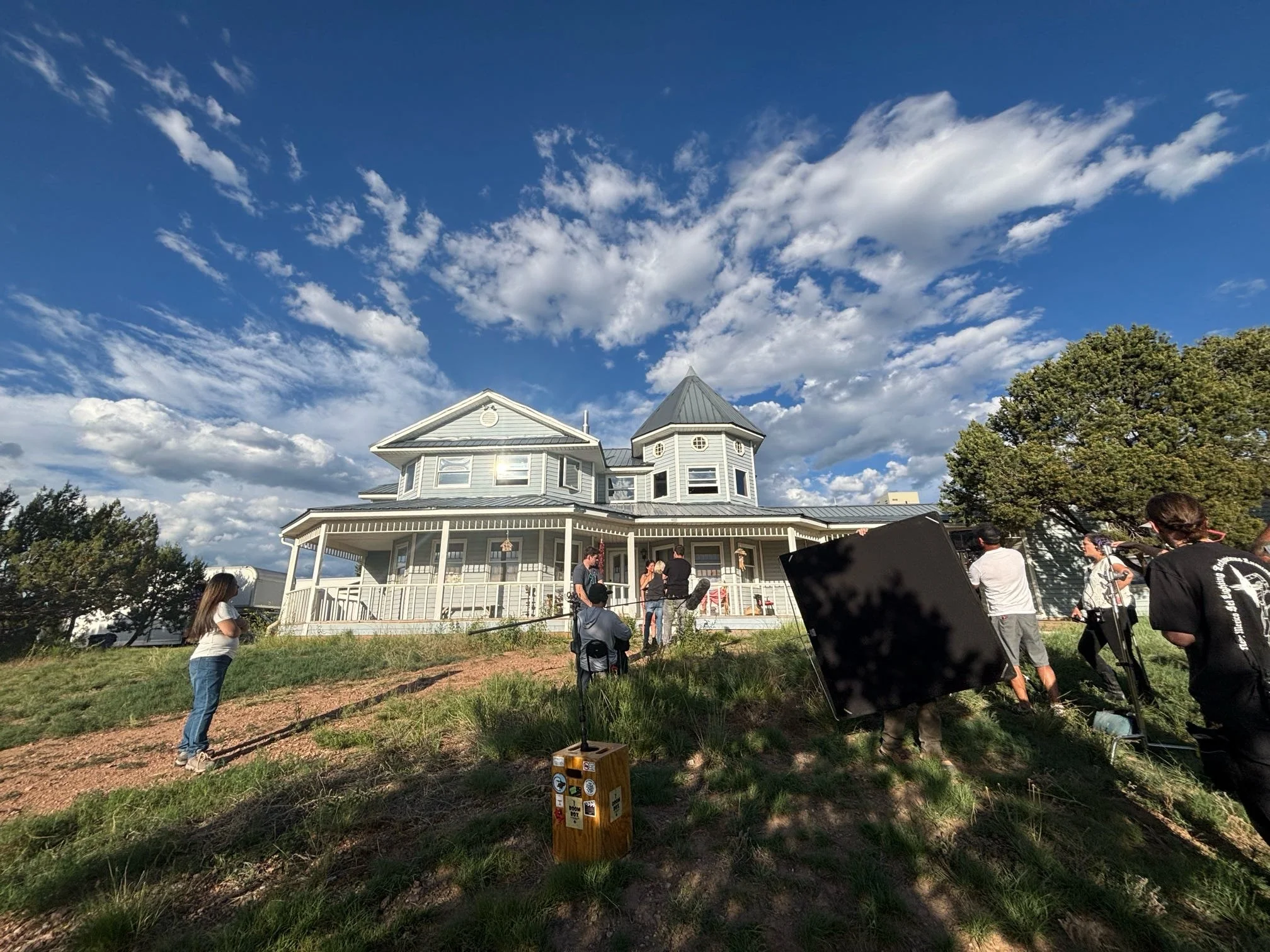Filming crew on a hill outside a large Victorian-style house with a blue sky and clouds overhead.