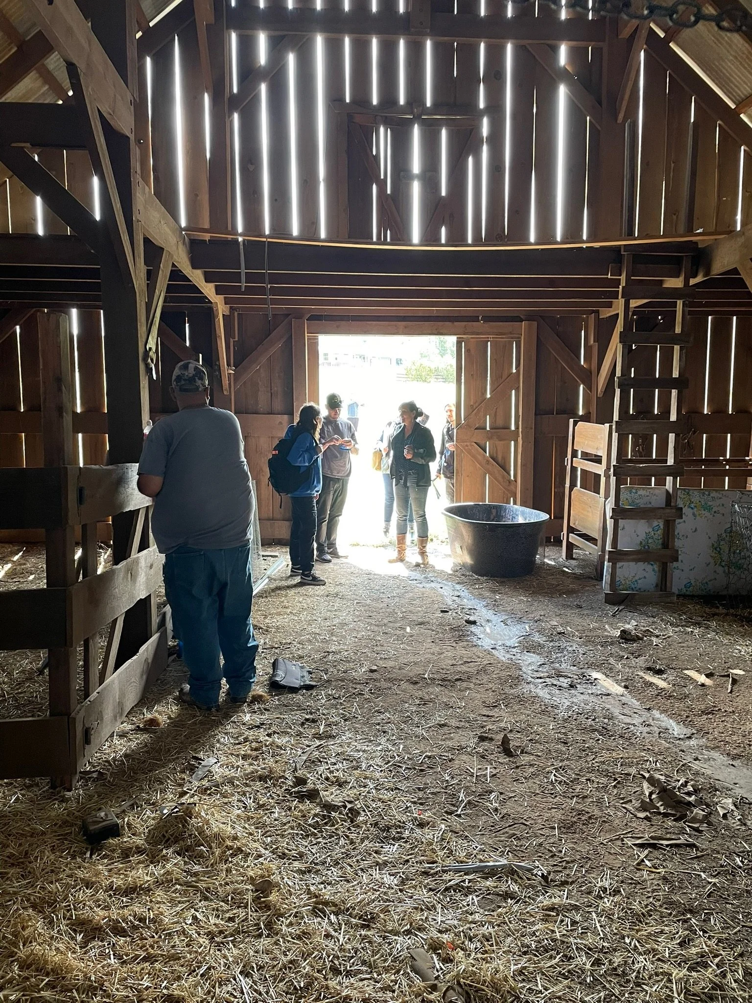 People gathered inside a rustic wooden barn near the entrance, with sunlight shining in. The barn has hay-straw on the floor, wooden beams, and an open door leading outside.