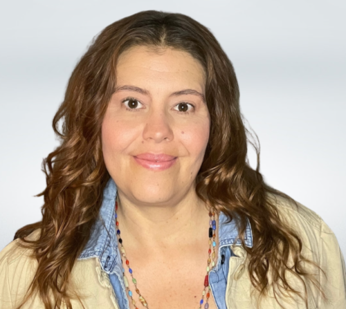 Portrait of a woman with curly brown hair, wearing a beige jacket over a denim shirt, and a colorful beaded necklace, smiling at the camera.