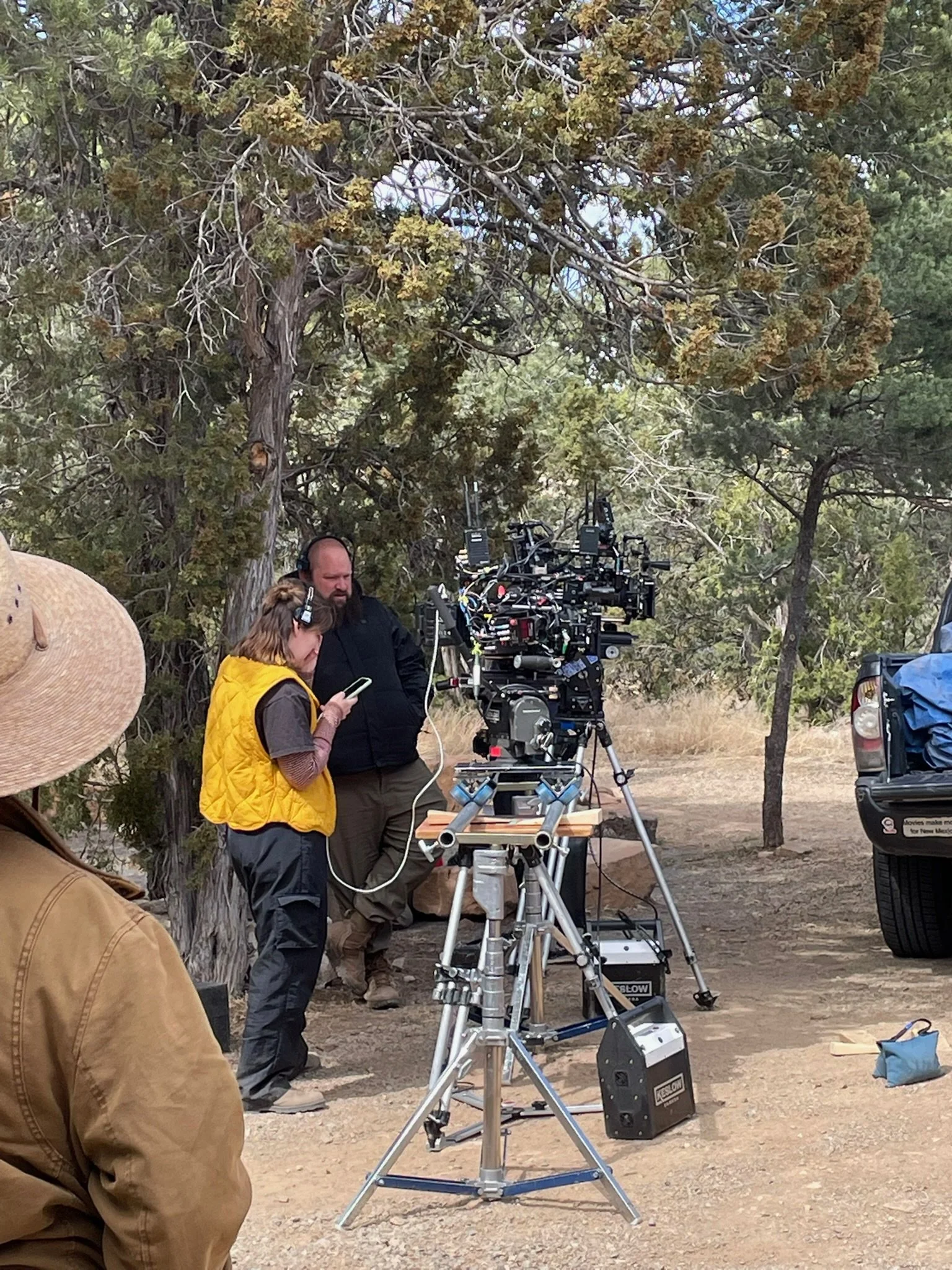 Film crew setting up camera equipment outdoors in a wooded area, with trees and a parked vehicle nearby.