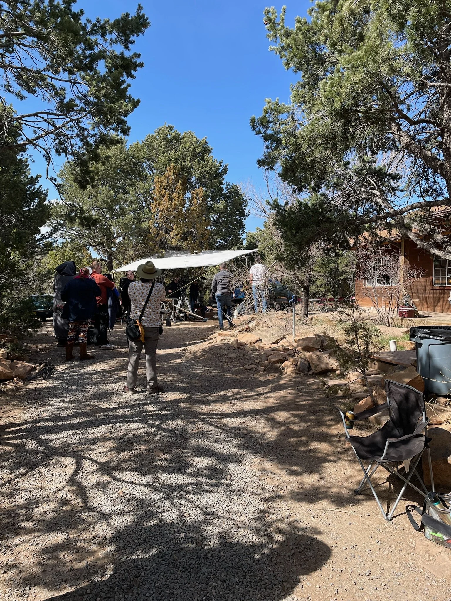 People gathered outdoors on a sunny day, standing under a shade cloth, surrounded by trees and rocks, near a wooden building, with folding chairs and a trash bin nearby.