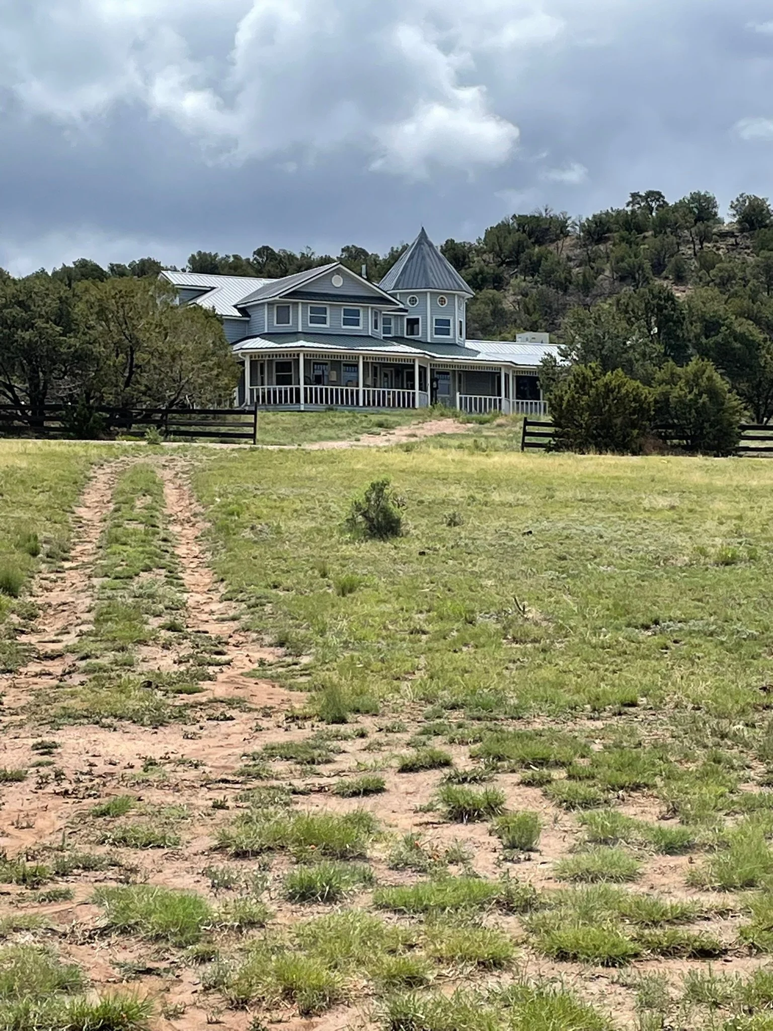 A large, blue, Victorian-style house with a turret sits on a hilltop on a cloudy day, surrounded by trees and grassy fields, with a dirt path leading up to it.