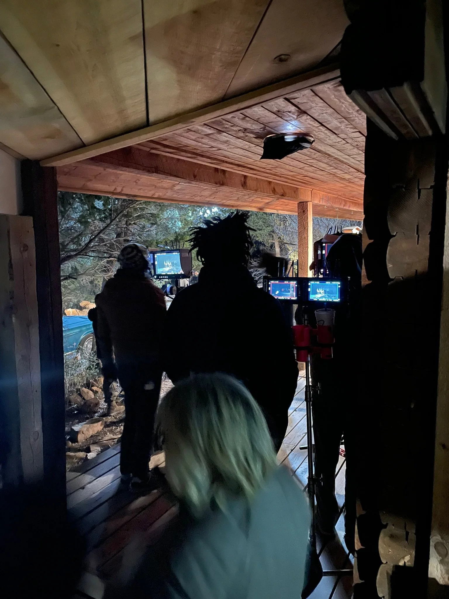 Filmmakers or videographers working with cameras and monitors on a porch with a wooded exterior in the background.