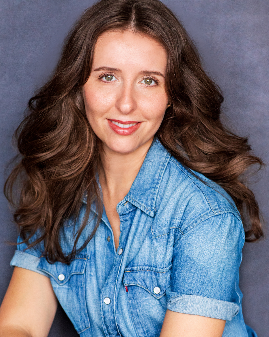 A young woman with long, wavy brown hair, wearing a denim shirt, smiling and looking at the camera against a dark background.