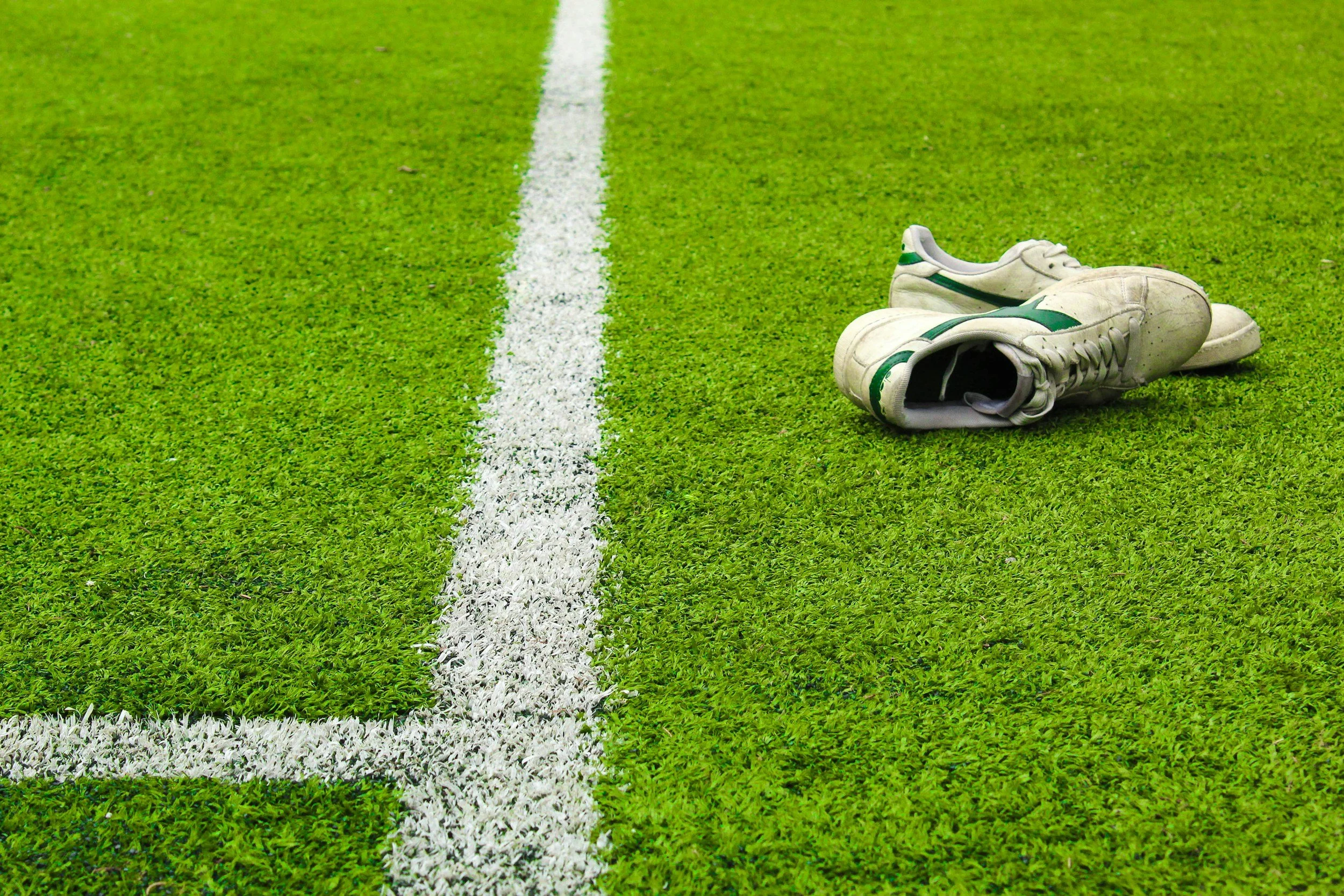 A pair of white and green soccer cleats lies on a green grass field next to a white line marking a soccer field boundary.