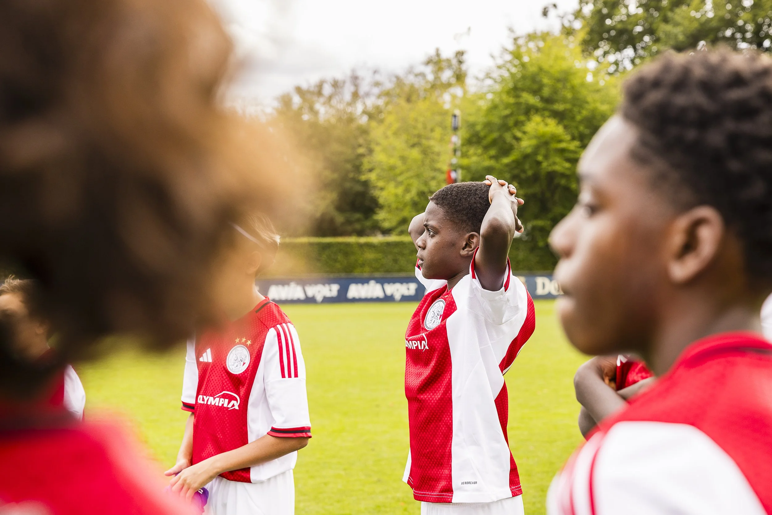 Young soccer players on of AJAX a grassy field, wearing red and white jerseys, with trees and a fence in the background. One boy in the center is holding his head behind his head, looking serious. During the Virgil's Legacy Trophy tournament in Tilbu