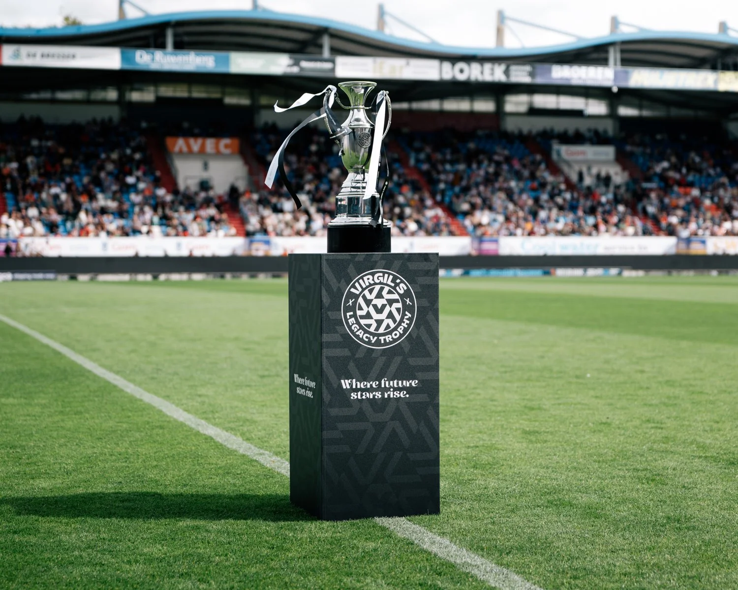 A trophy on a black pedestal with the logo and text for Virgili's Legacy Trophy, on a soccer field with a stadium and crowd in the background.