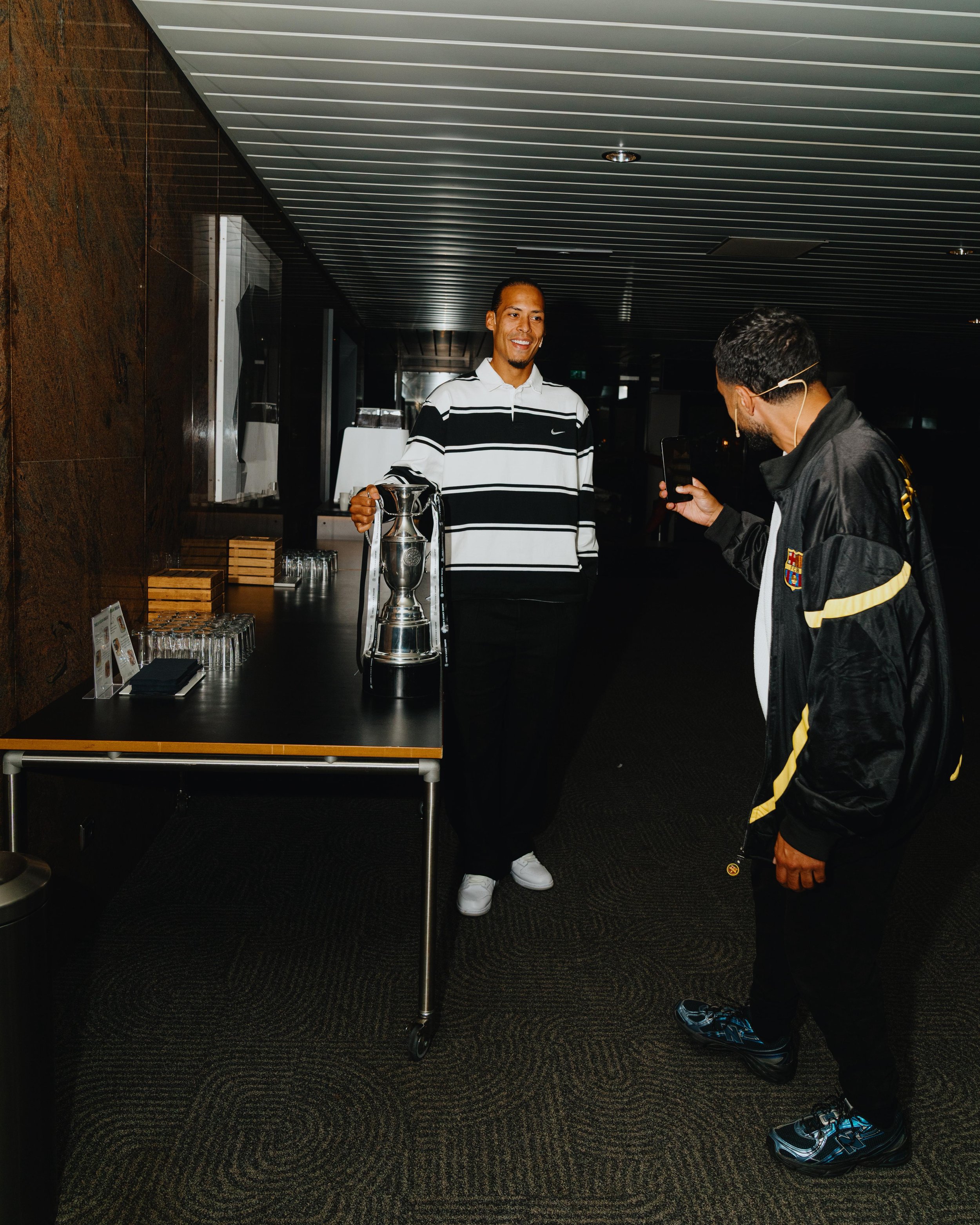 A man in a black and white striped shirt holding a large silver trophy, smiling, is being photographed by another man in a black jacket with yellow accents. They are in a dimly lit indoor space with a table that has glasses and boxes on it. During th