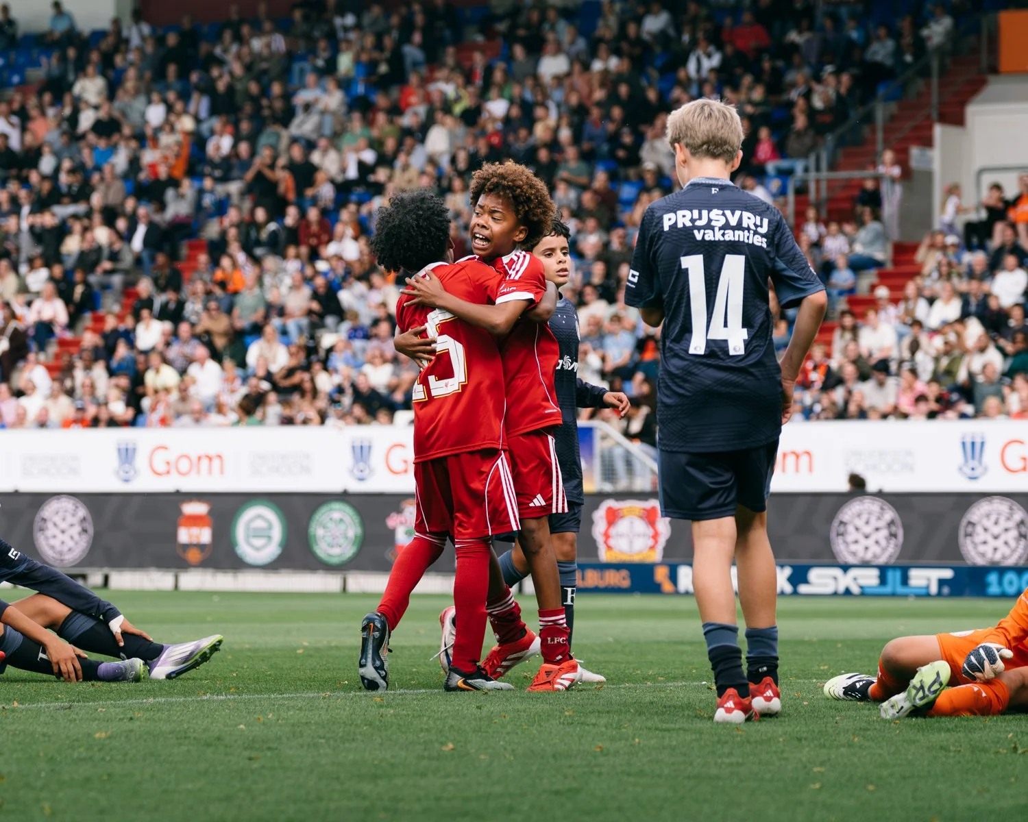 Young soccer players, some wearing red uniforms, are emotional and comforting each other on the field during a game, with spectators in the stands watching.