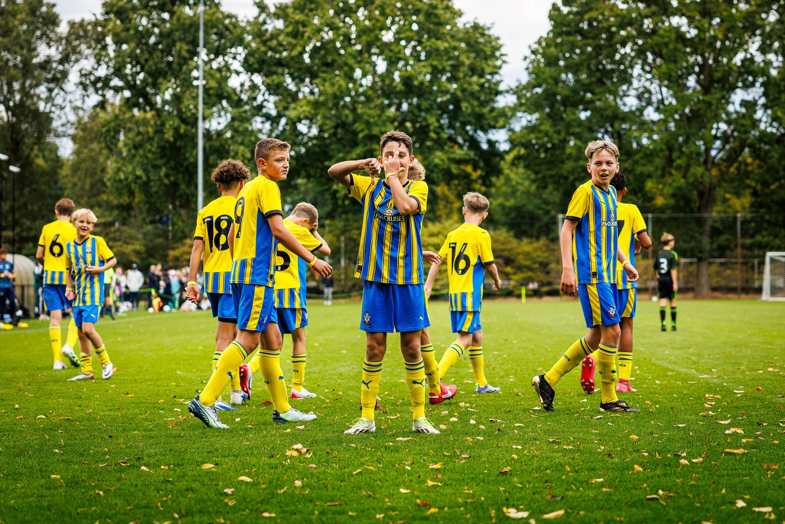 Children playing soccer on a green field, wearing yellow and blue uniforms with some players standing and one making a gesture, with trees and spectators in the background. During the Virgil's Legacy Trophy tournament in Tilburg.