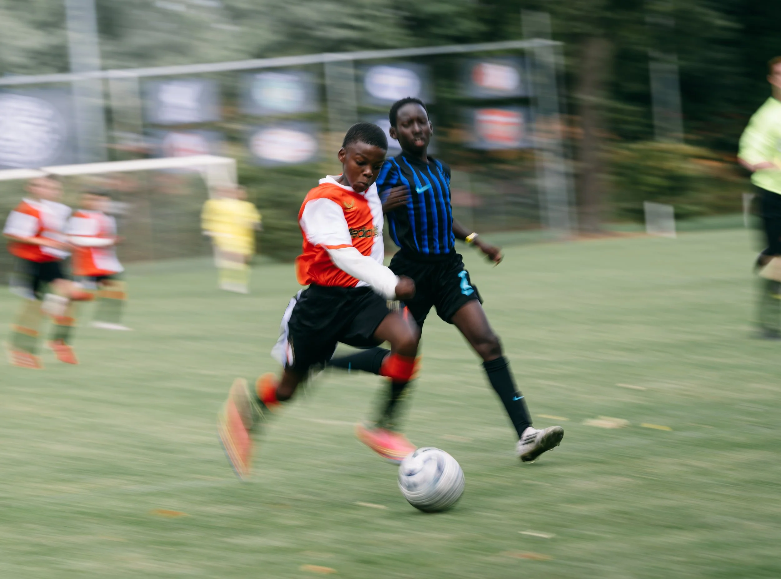 Two young boys playing soccer on a grassy field, with other children and players in the background. One boy in an orange and white jersey is running towards a soccer ball while the other in a blue and black jersey is nearby. The background is blurred, indicating movement.