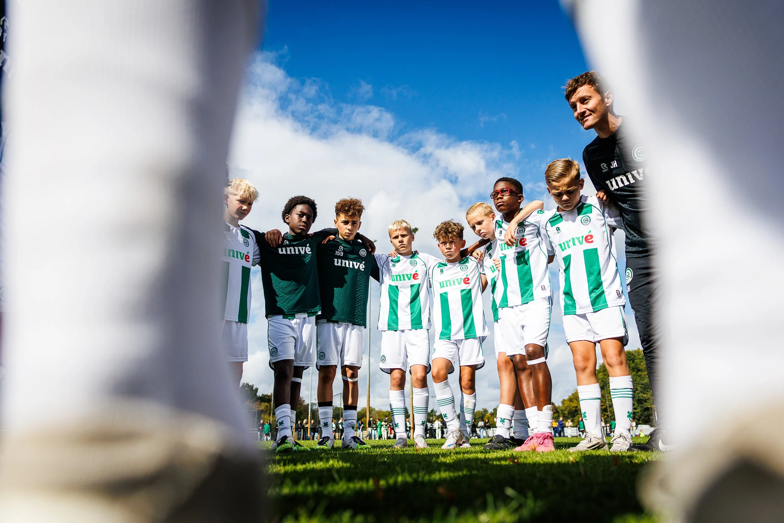 Young soccer players in green and white uniforms listen to coach during team huddle on grassy field, viewed from behind goal net. During the Virgil's Legacy Trophy tournament in Tilburg.