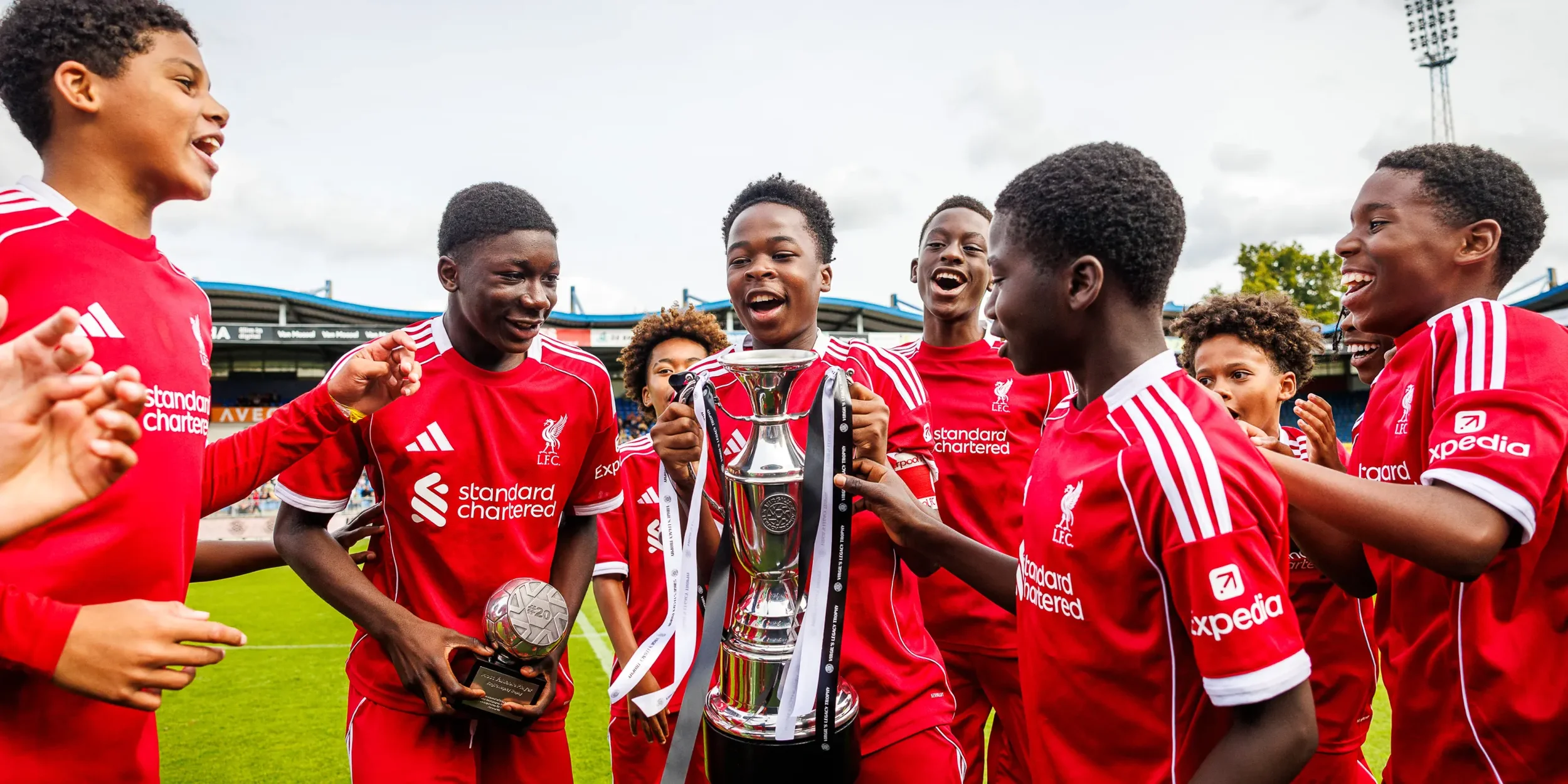 A group of young soccer players in red jerseys celebrating on a soccer field, holding a large trophy, with some holding medals, smiling and cheering.