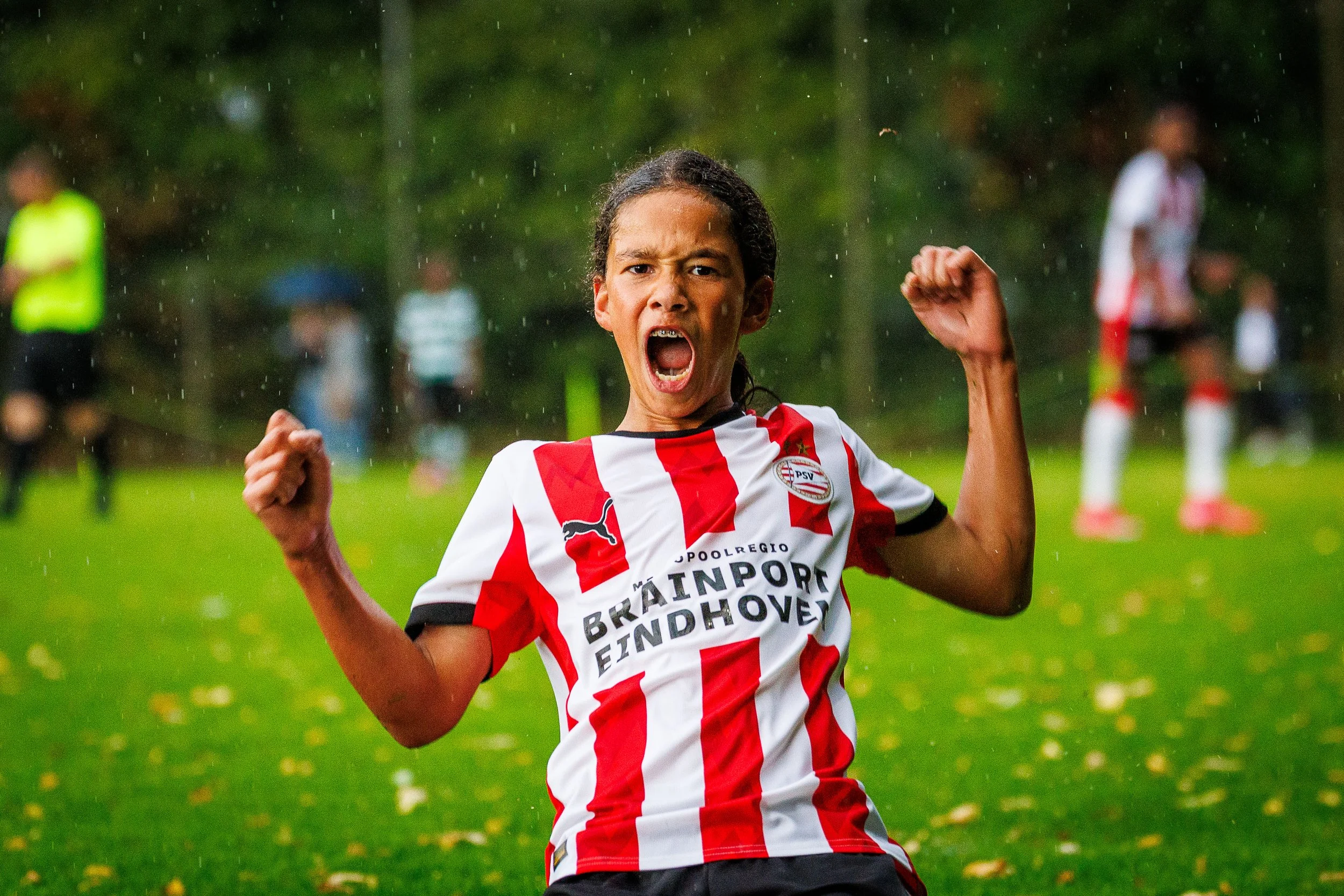 Young player celebrating a goal, wearing a red and white striped soccer jersey, with a fierce expression and clenched fists, on a rainy day on the soccer field. During the Virgil's Legacy Trophy tournament in Tilburg.