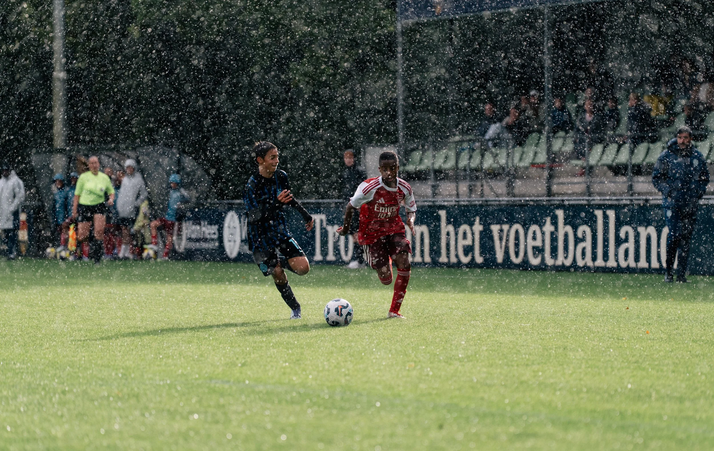 Two young boys playing soccer in the rain on a grass field, with spectators in the background and a large signboard.
