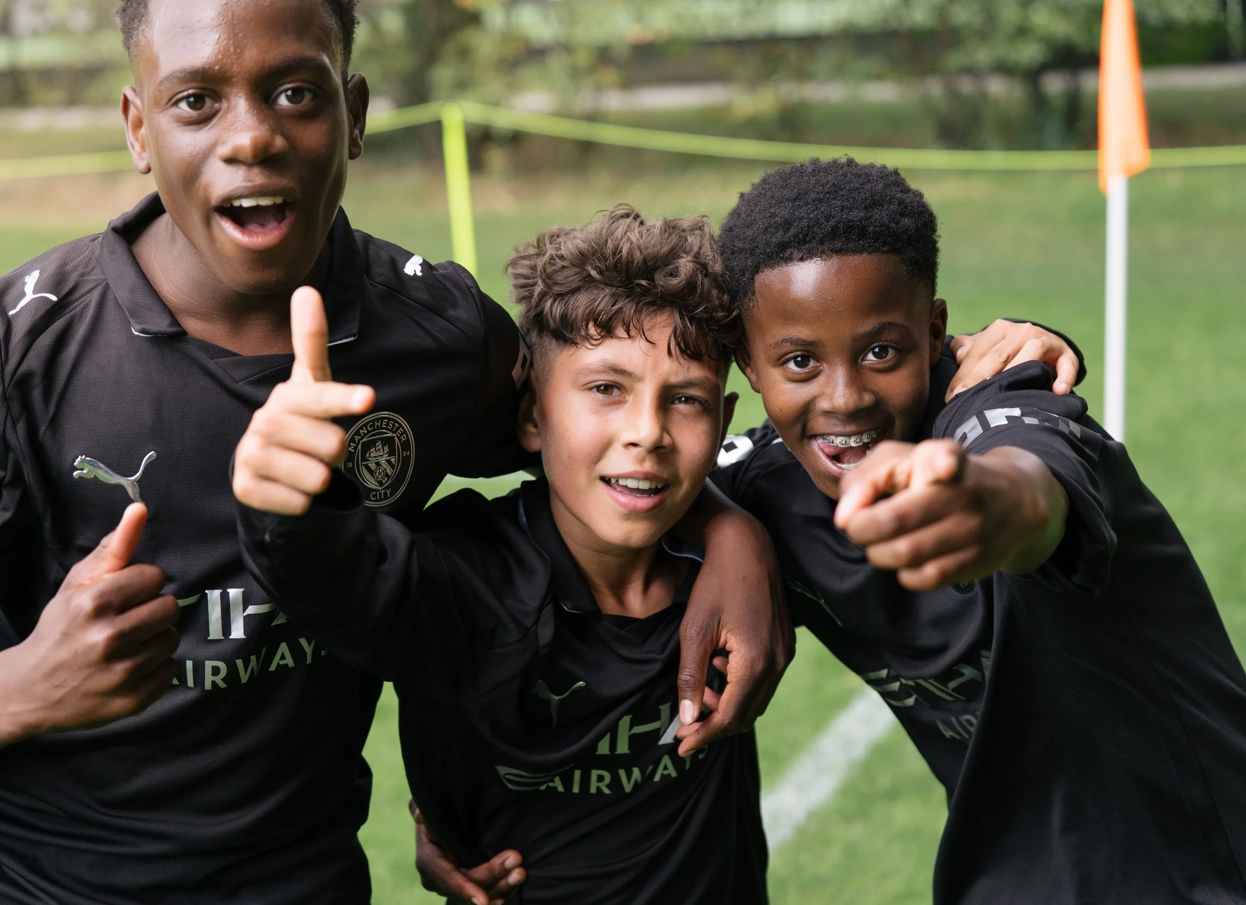 Three boys in black football jerseys of Manchester City posing together on a soccer field, smiling and pointing at the camera, with a grassy field and soccer goal in the background. During the Virgil's Legacy Trophy tournament in Tilburg.