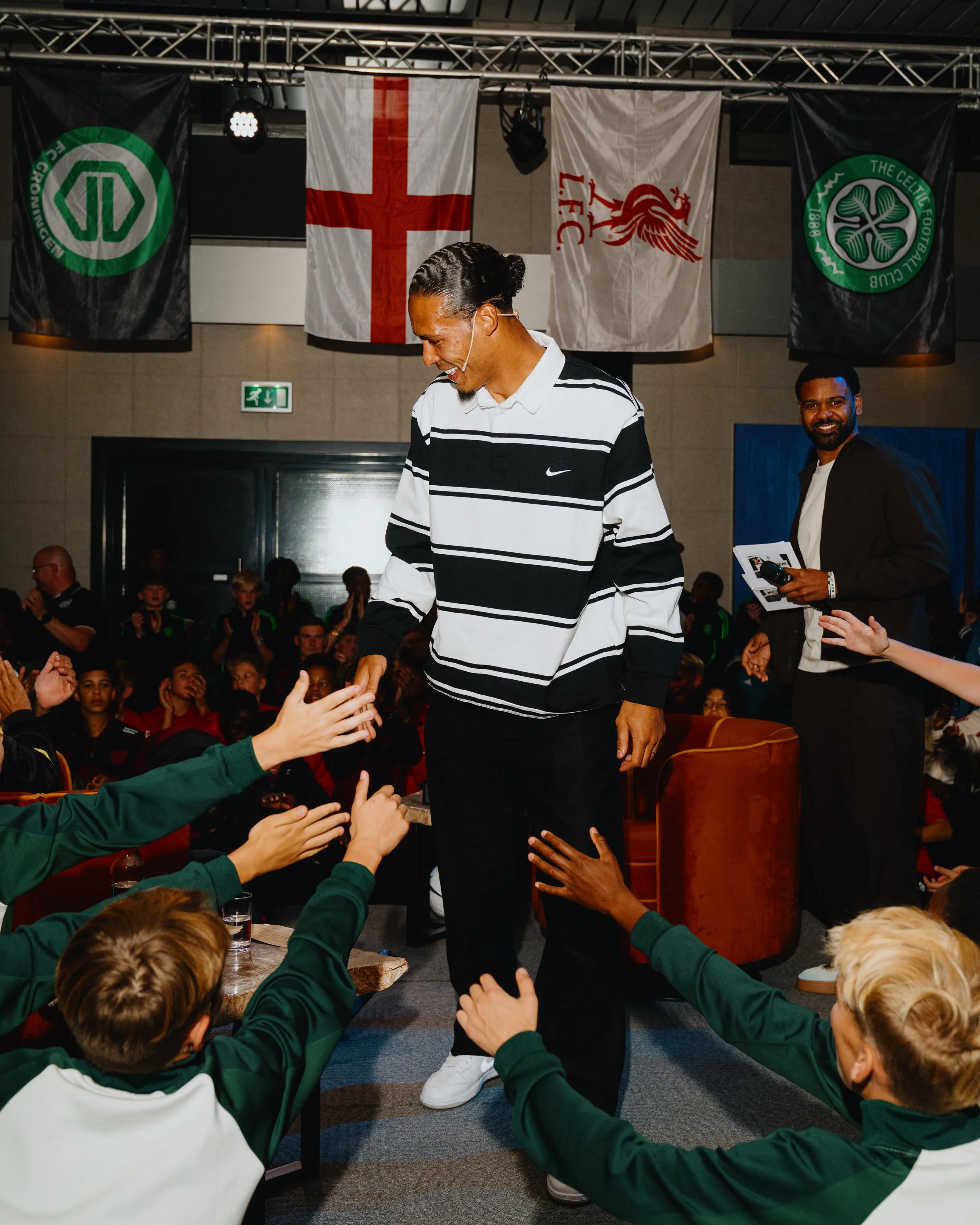 Virgil van Dijk in a black and white striped Nike shirt shaking hands with children at an event, with a speaker standing behind him and flags hanging in the background. During the Virgil's Legacy Trophy tournament in Tilburg.