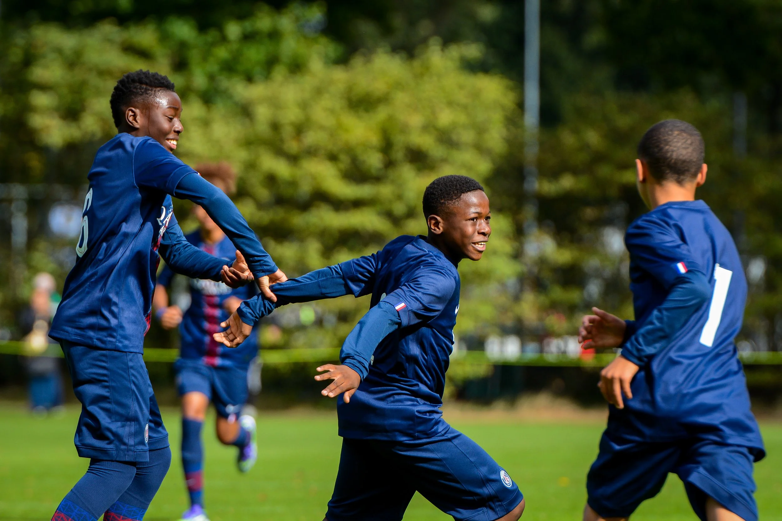 Three boys playing soccer on a green field during daytime, wearing navy blue uniforms, with trees in the background. During the Virgil's Legacy Trophy tournament in Tilburg.
