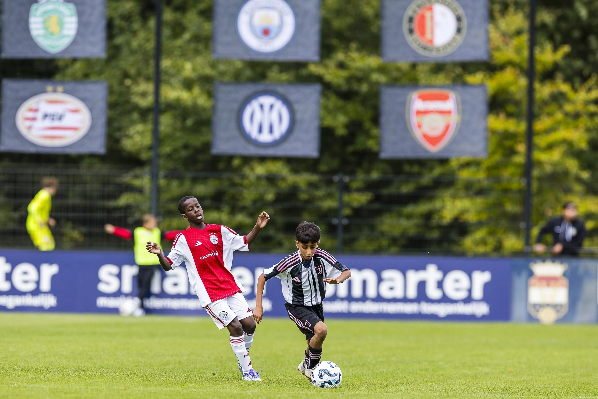 Two boys playing soccer on a grass field during a match, with one in a red and white jersey and the other in a black and white striped jersey. Several flags representing different soccer clubs hang in the background.